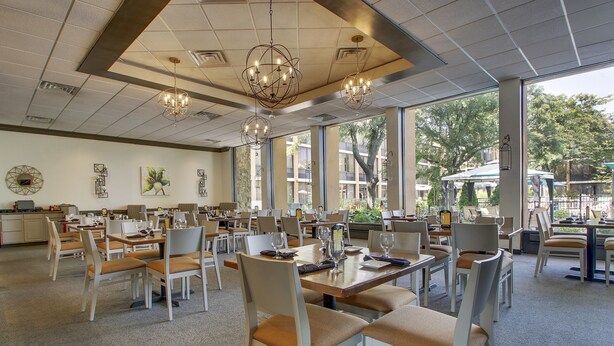 A spacious dining area with multiple tables and chairs, large windows offering a view of the outdoors, and decorative lighting fixtures. - Holiday Inn Westbury - Long Island by IHG (Carle Place)