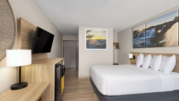 A hotel room with a bed, a desk with a TV, a wall-mounted picture, and a door in the background.