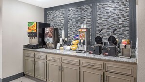 A self-service breakfast bar with various food and drink options, including a coffee machine, waffle maker, and a beverage dispenser.