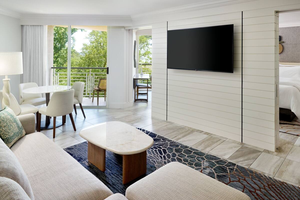 Ein Hotelzimmer mit großem Flachbildfernseher an der Wand, einer Sitzecke mit Sofa, einem Couchtisch sowie einem Essbereich mit Stühlen und Tisch.