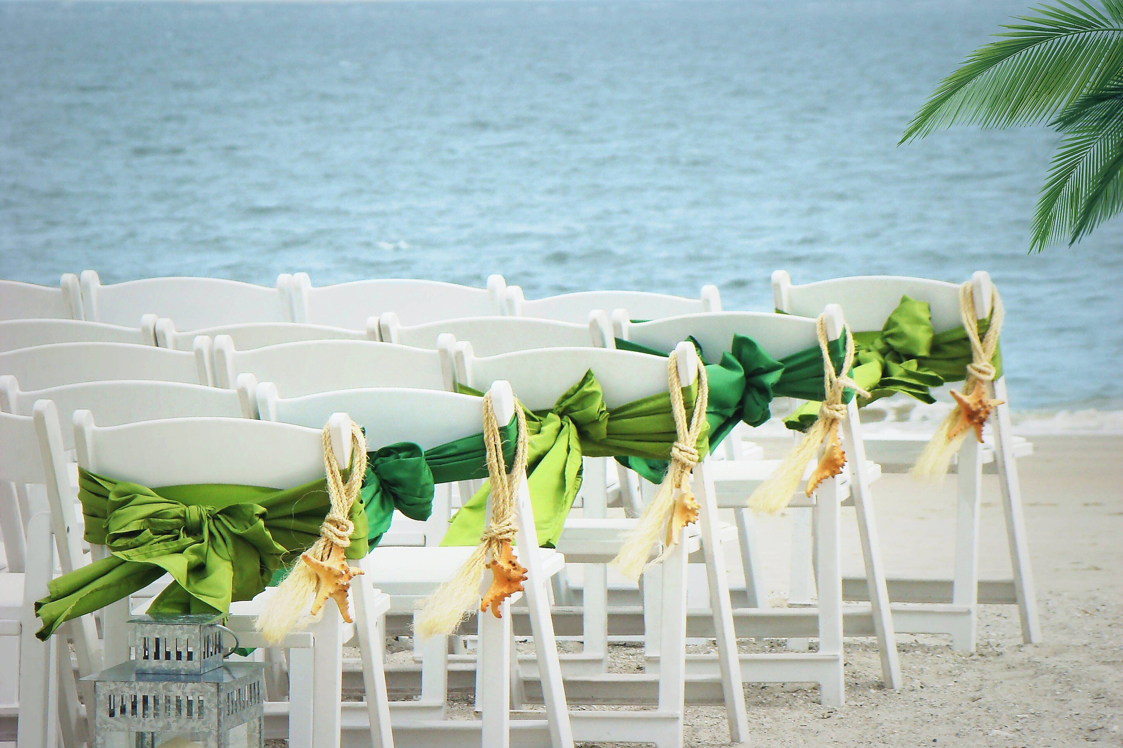 White plastic chairs with green and brown decorations, arranged on a sandy beach with a view of the ocean.