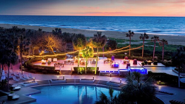 Une piscine d’un complexe hôtelier offrant une vue sur l’océan au coucher du soleil, entourée de palmiers et illuminée de guirlandes lumineuses.