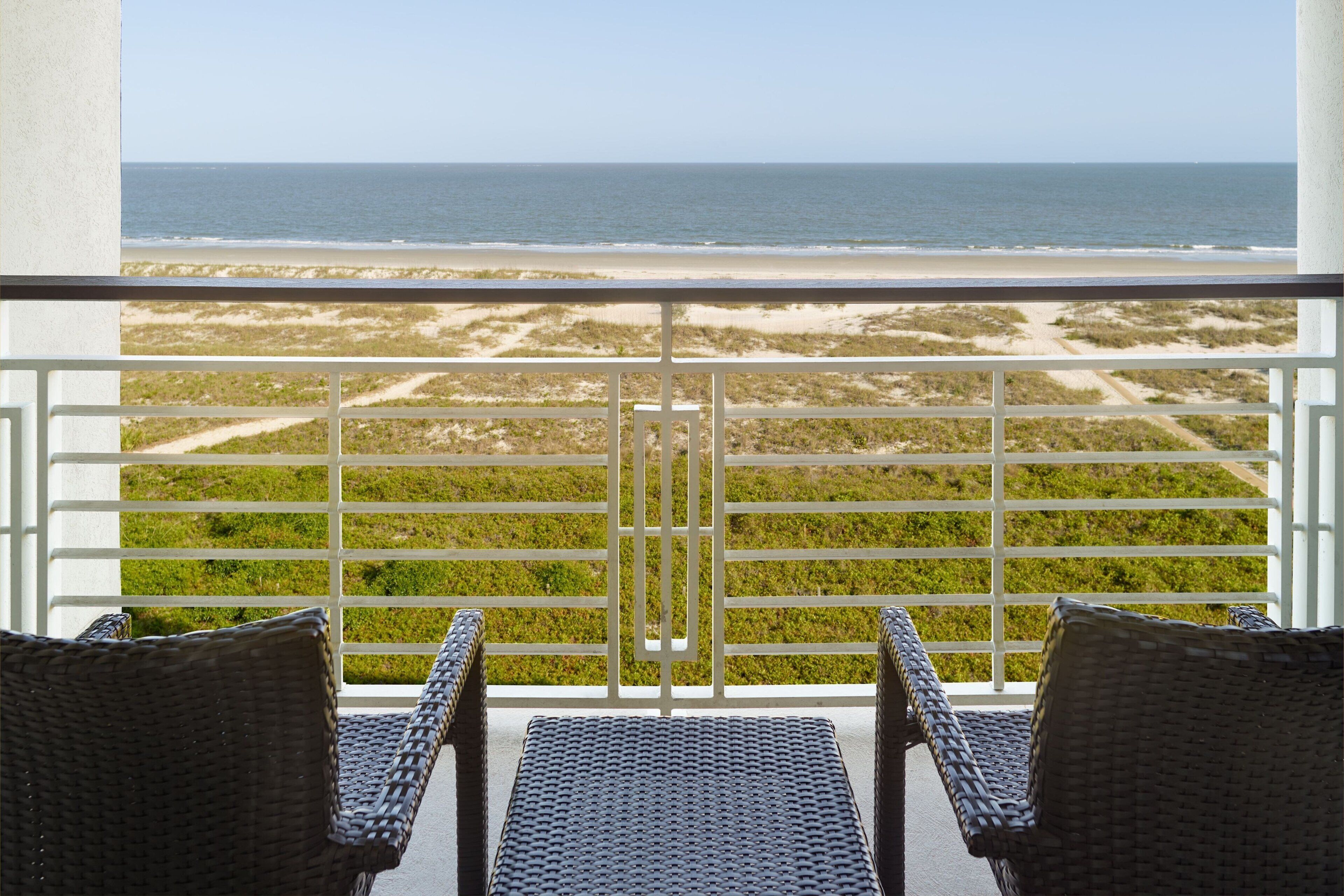 Ein Balkon mit Korbstühlen mit Blick auf einen Strand mit Sand und spärlicher Vegetation.