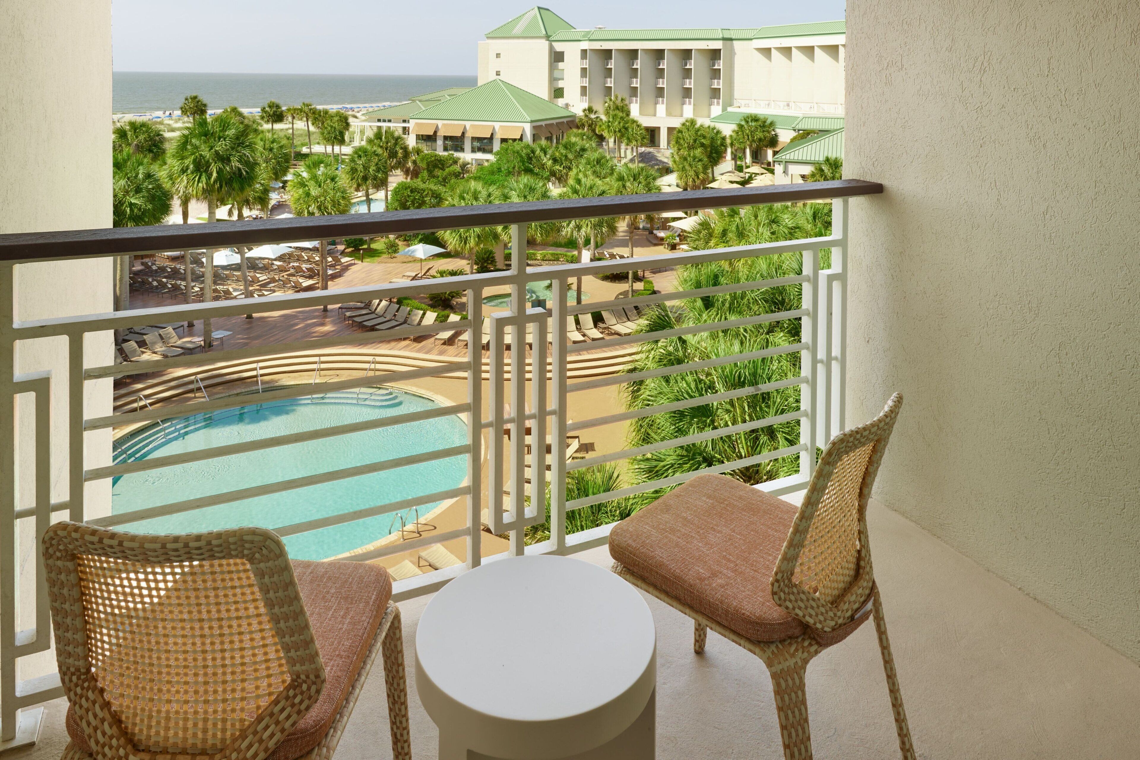 A balcony with wicker chairs and a small table, overlooking a pool and a resort area with palm trees and a building in the background.