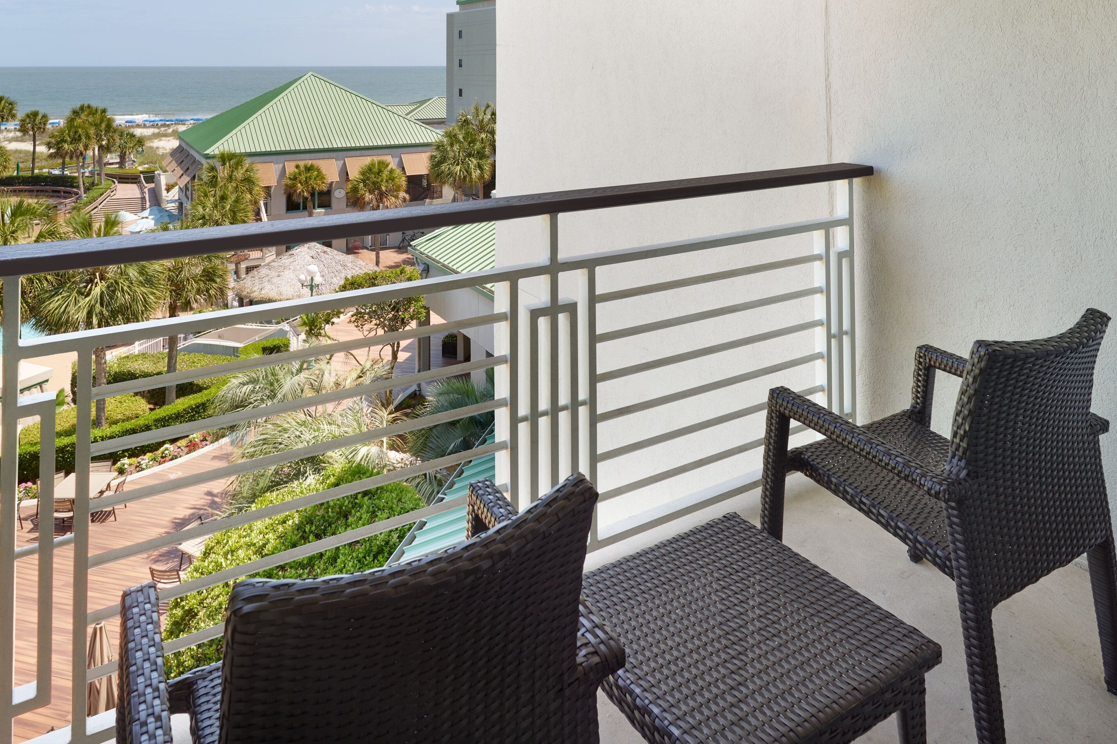 A balcony with wicker furniture overlooking a resort area with a pool, palm trees, and a view of the ocean.