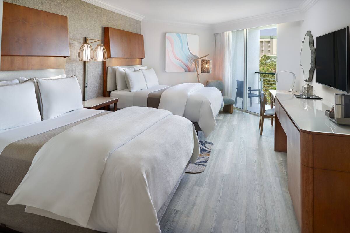 Ein Hotelzimmer mit zwei Betten, einem Schreibtisch, einem Sessel, einem Fernseher und einem Balkon mit Aussicht.