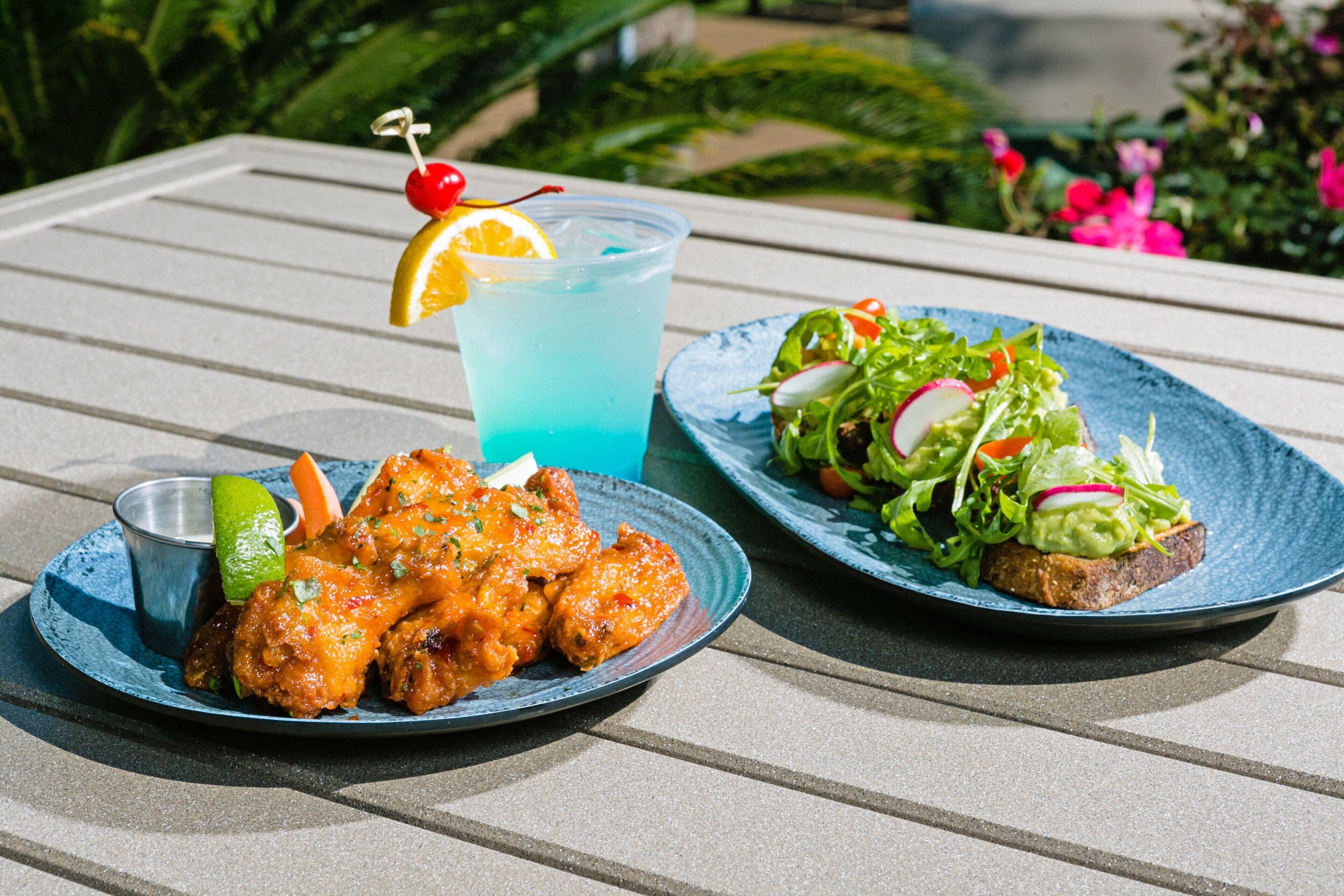 A table with a plate of chicken wings, a salad, and a blue drink with a cherry and orange slice garnish.
