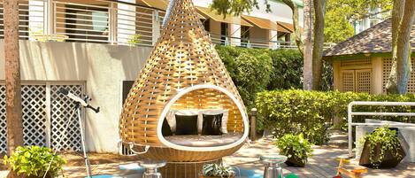 A hanging chair with a circular seat and a conical roof, surrounded by potted plants and outdoor furniture.