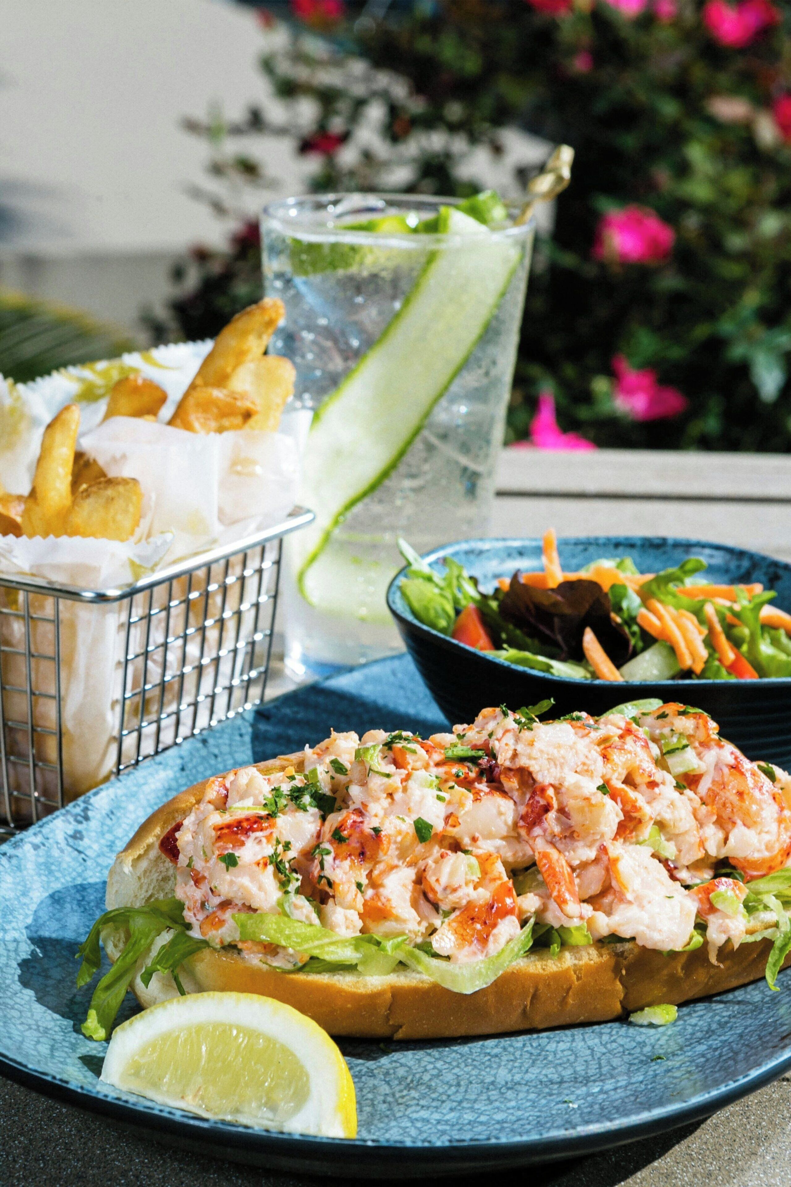 A seafood sandwich on a blue plate, a side salad, a glass of iced drink, and a basket of fries.