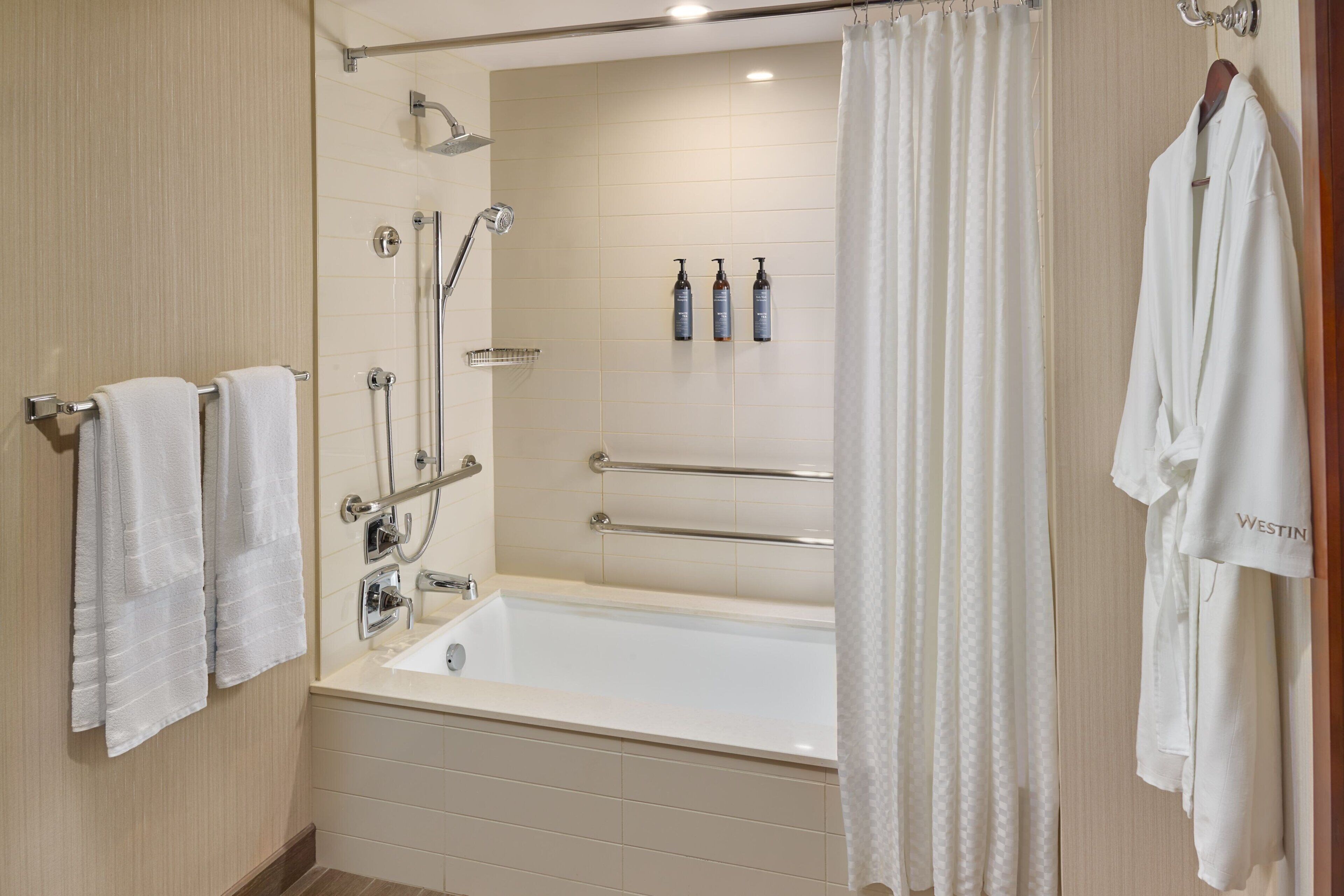 A modern bathroom with a bathtub, shower, and handrails. There are towels and bottles on the wall.