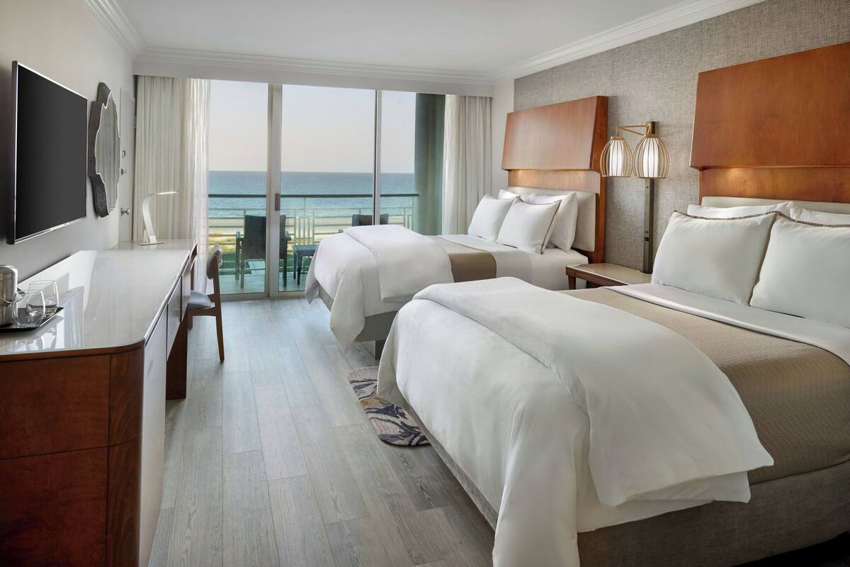 Ein Hotelzimmer mit zwei Betten, einem Schreibtisch, einem Fernseher und einem Balkon mit Meerblick.