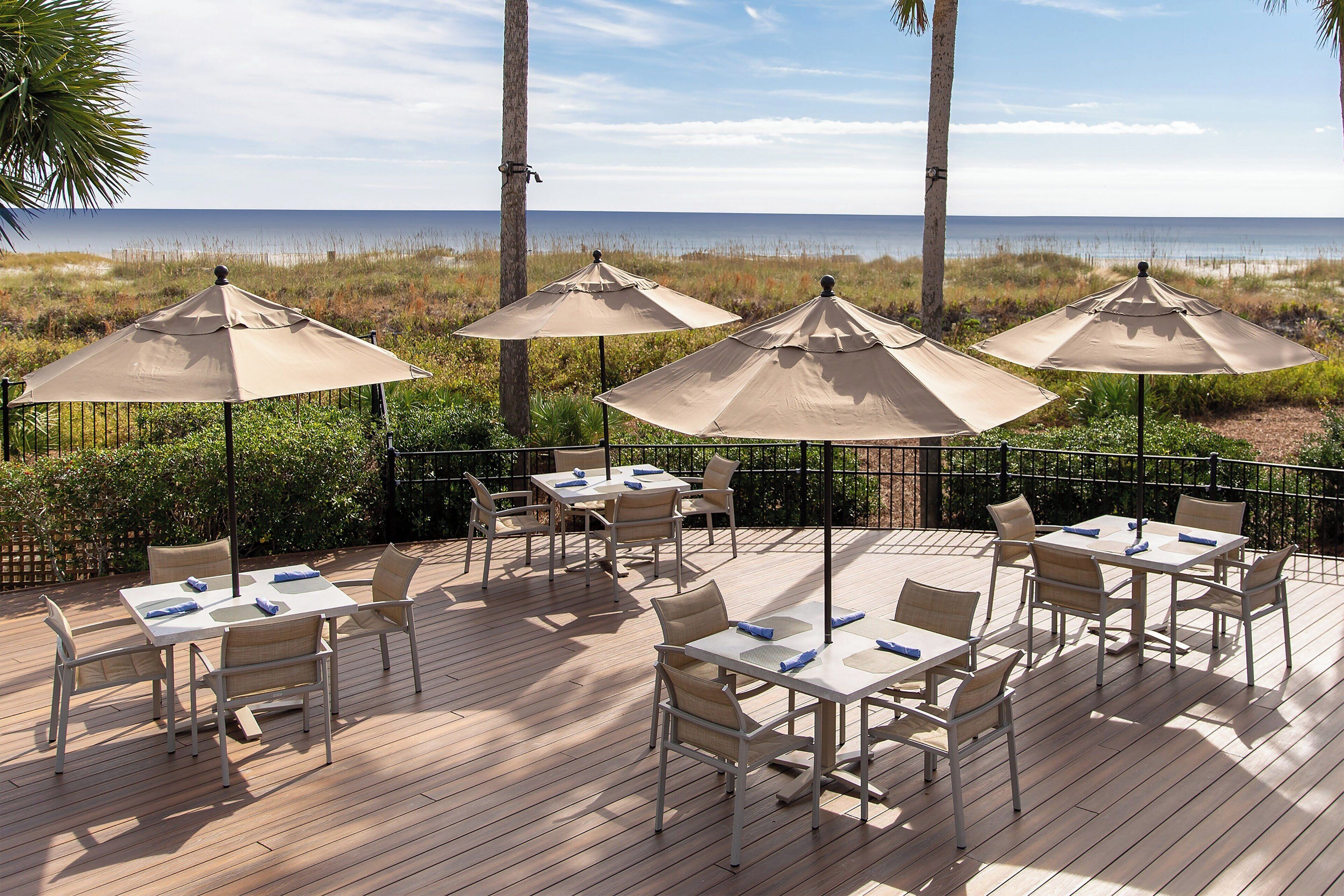 A deck with tables and chairs, beige umbrellas, and a view of the ocean.