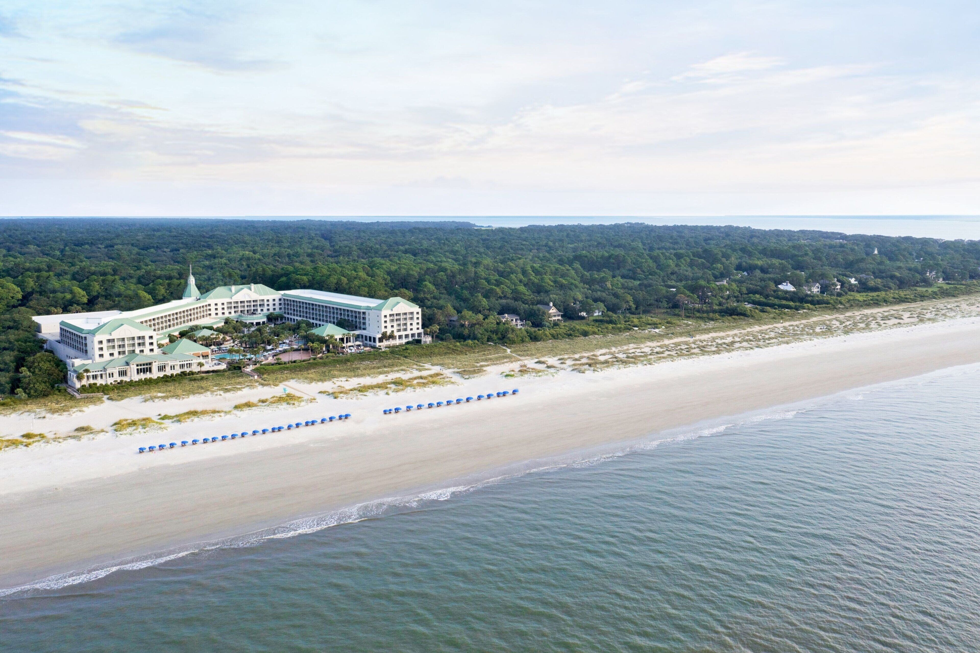 Un grand hôtel de villégiature comprenant un bâtiment central et plusieurs ailes, situé sur une plage avec vue sur l’océan et des collines boisées en arrière-plan.