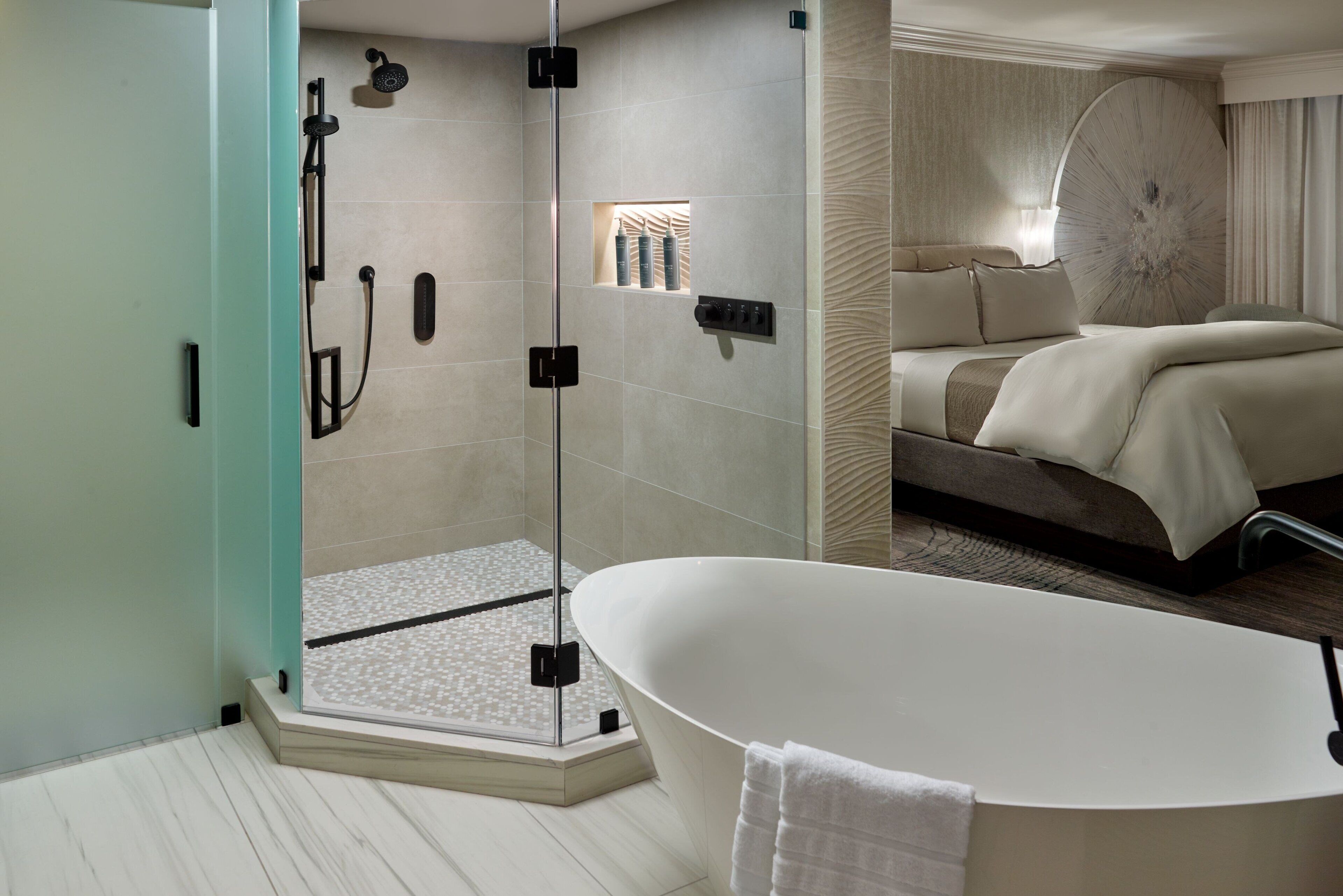 A modern bathroom with a glass-enclosed shower, a freestanding bathtub, and a bed with a large mirror and curtains.