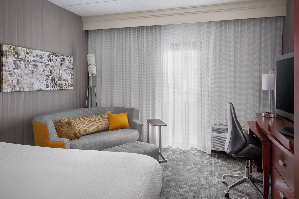 A hotel room with a bed, a sofa, a desk, a TV, and a large window with curtains.
