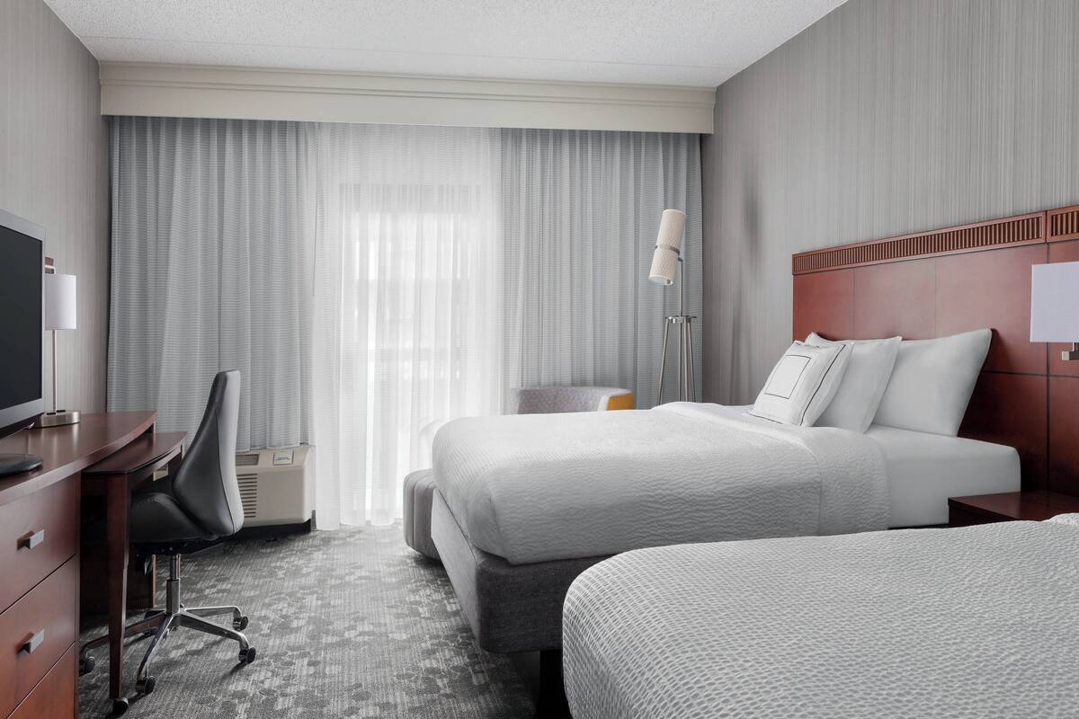 A hotel room with two beds, a desk, a chair, a TV, and a lamp.