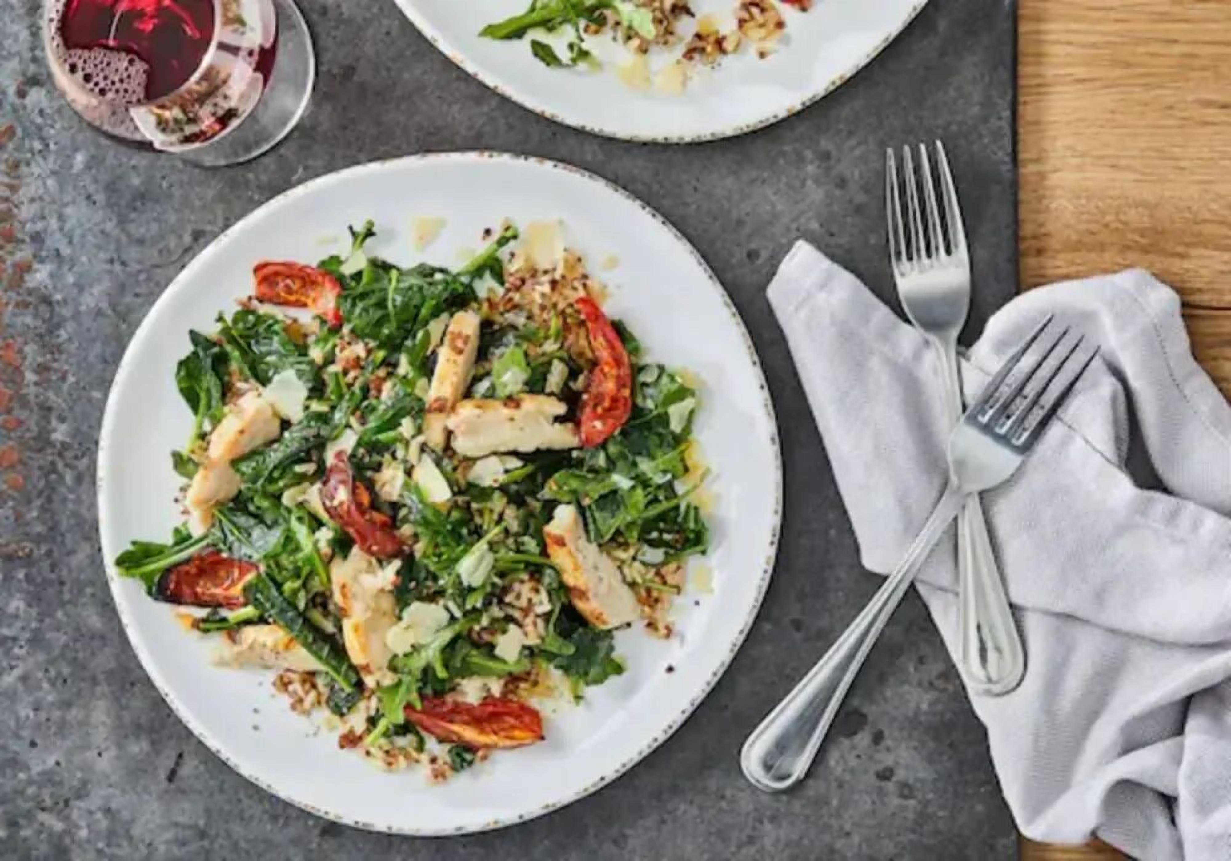 A plate of food with greens, tomatoes, and cheese on a table with a fork and napkin.