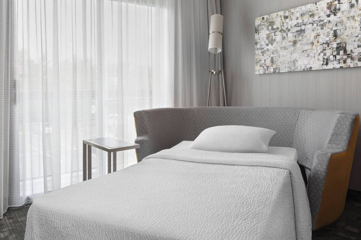 A modern hotel room with a large bed, a small side table, a floor lamp, and a textured abstract painting on the wall.