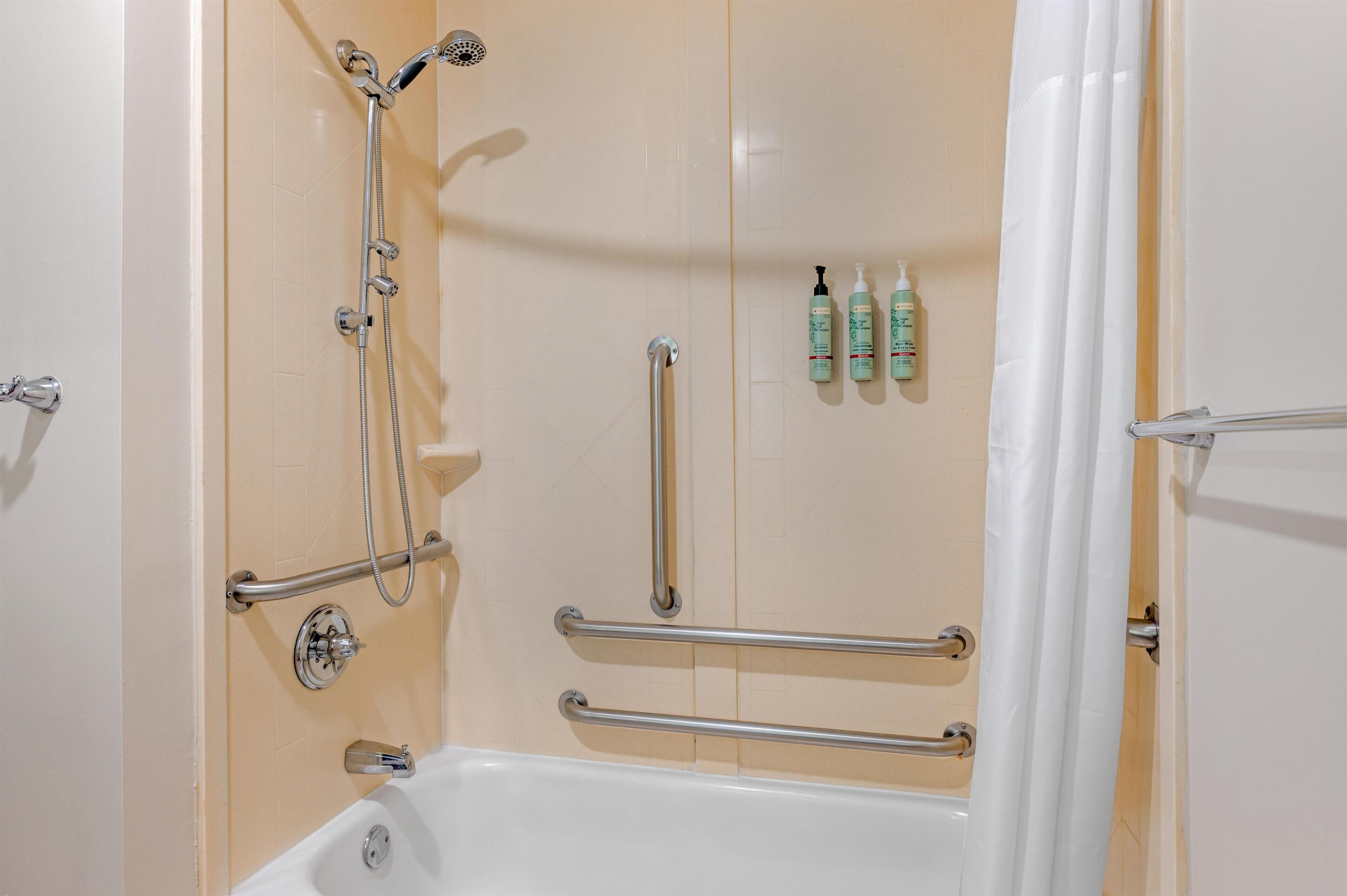 A bathroom with a bathtub, shower, grab bars, and toiletries on the wall.