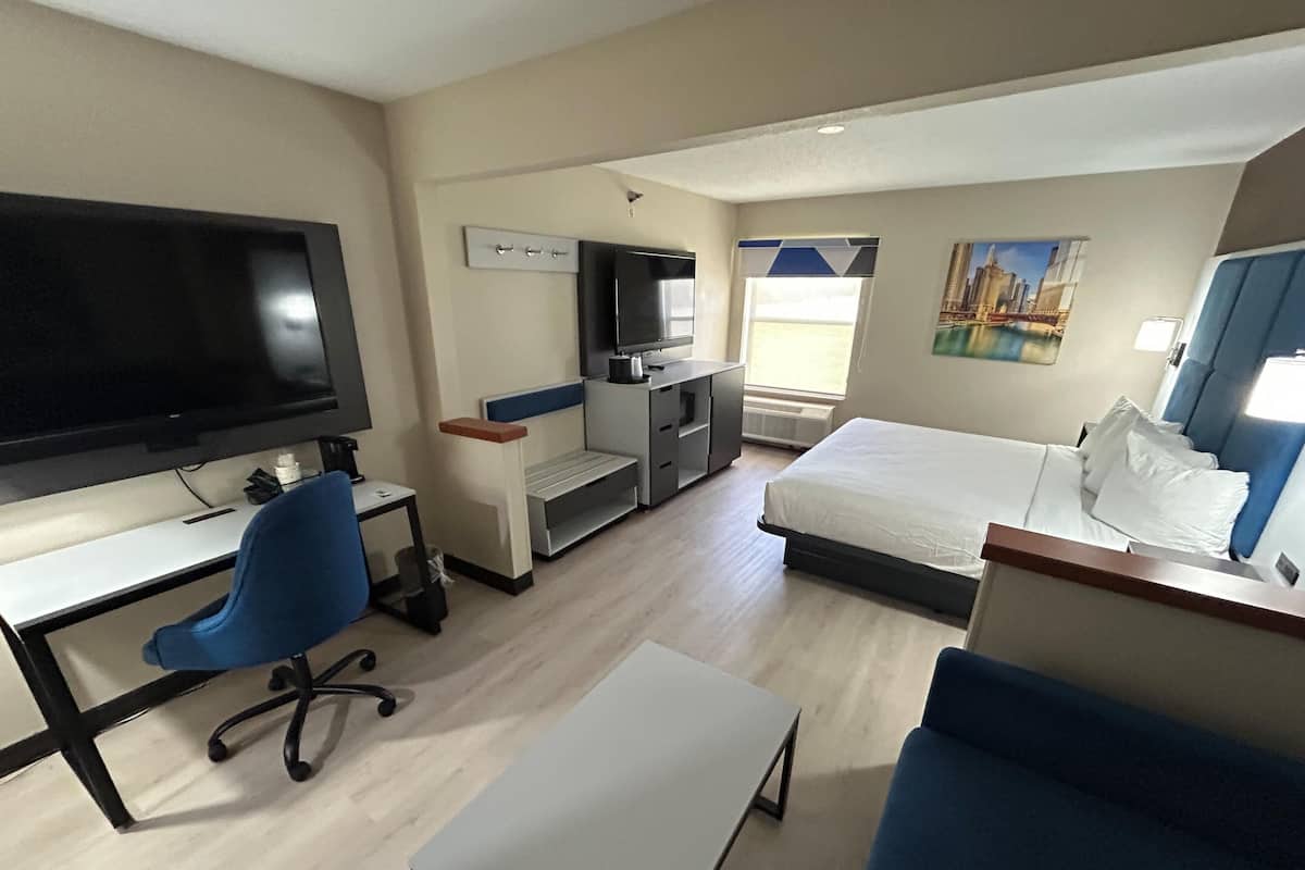 A hotel room with a bed, a desk with a chair, a TV, a small kitchenette, and a window with a view.