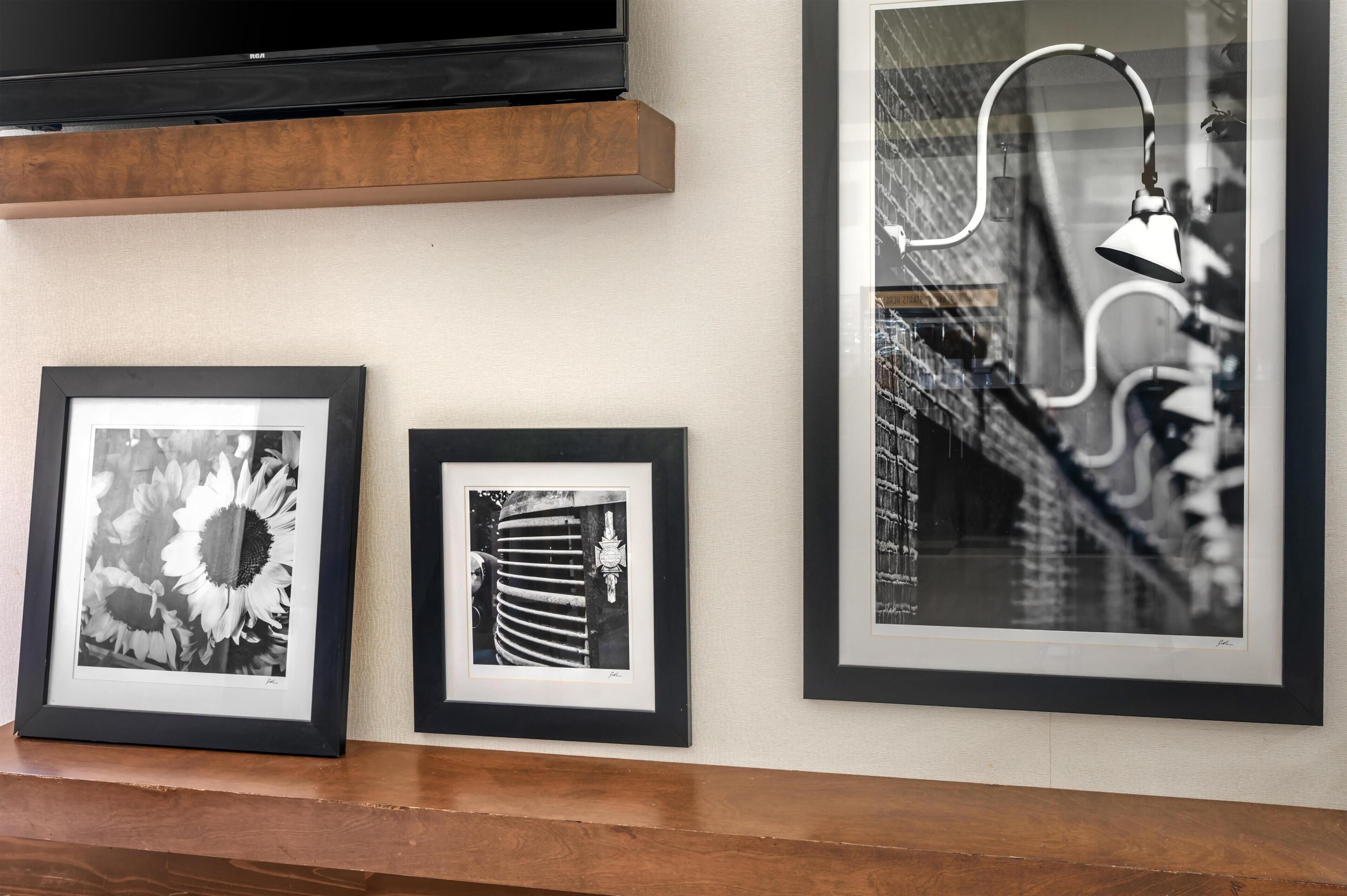 A wall with three framed black and white photographs, a television above, and a wooden shelf below.