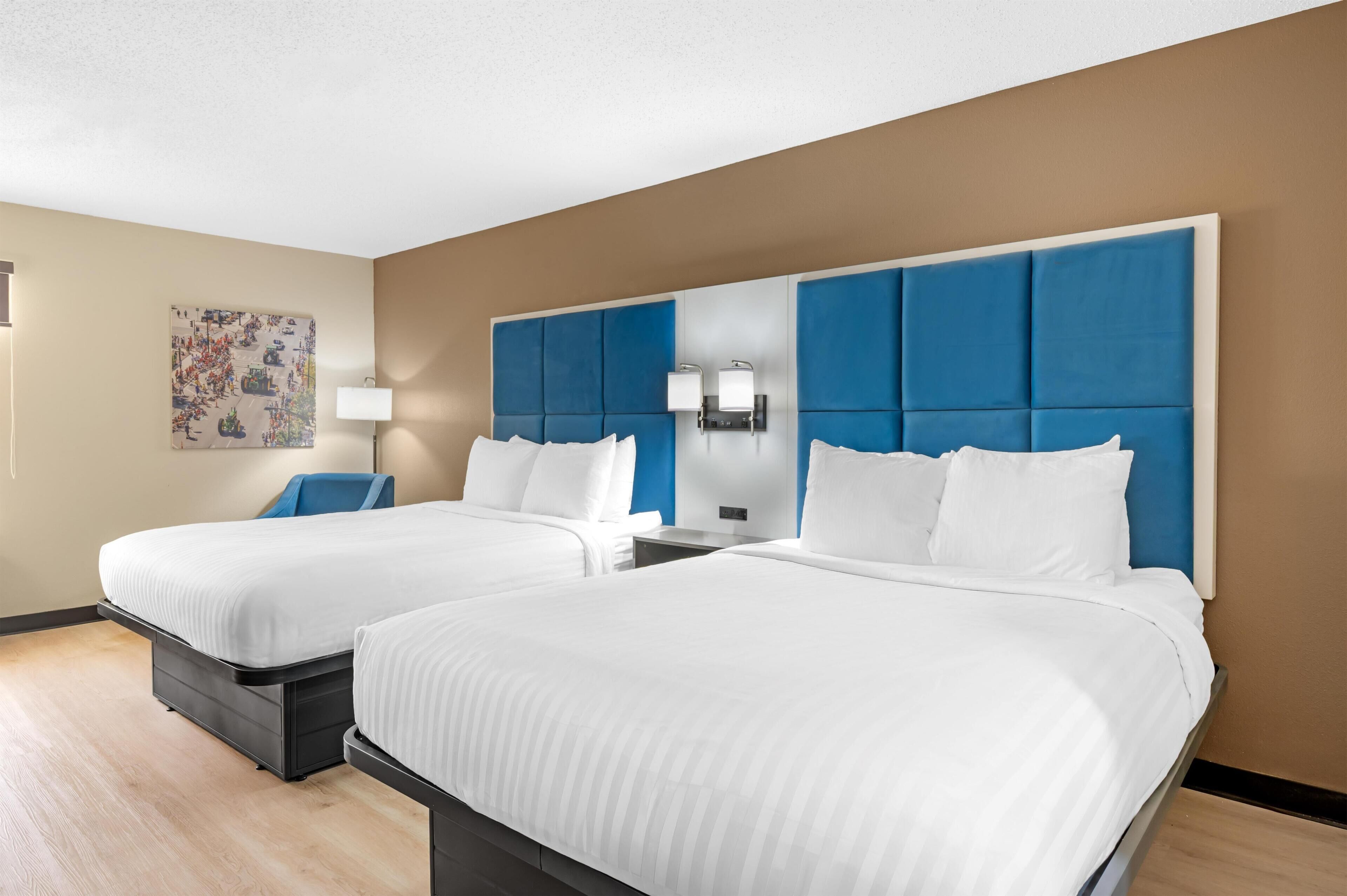A hotel room with two beds, a blue headboard, a wall-mounted lamp, and a painting on the wall.