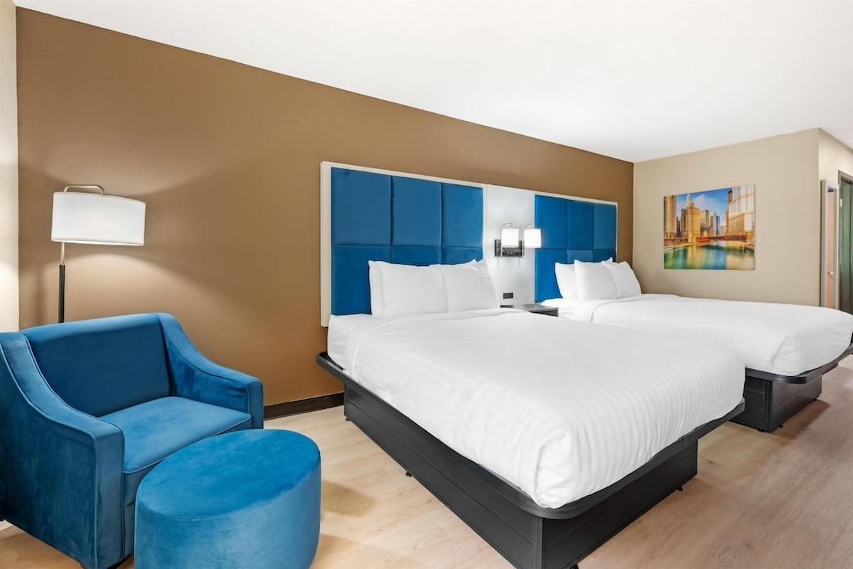 Een hotelkamer met een tweepersoonsbed, een eenpersoonsbed, een blauwe fauteuil, een blauwe poef en een lamp.