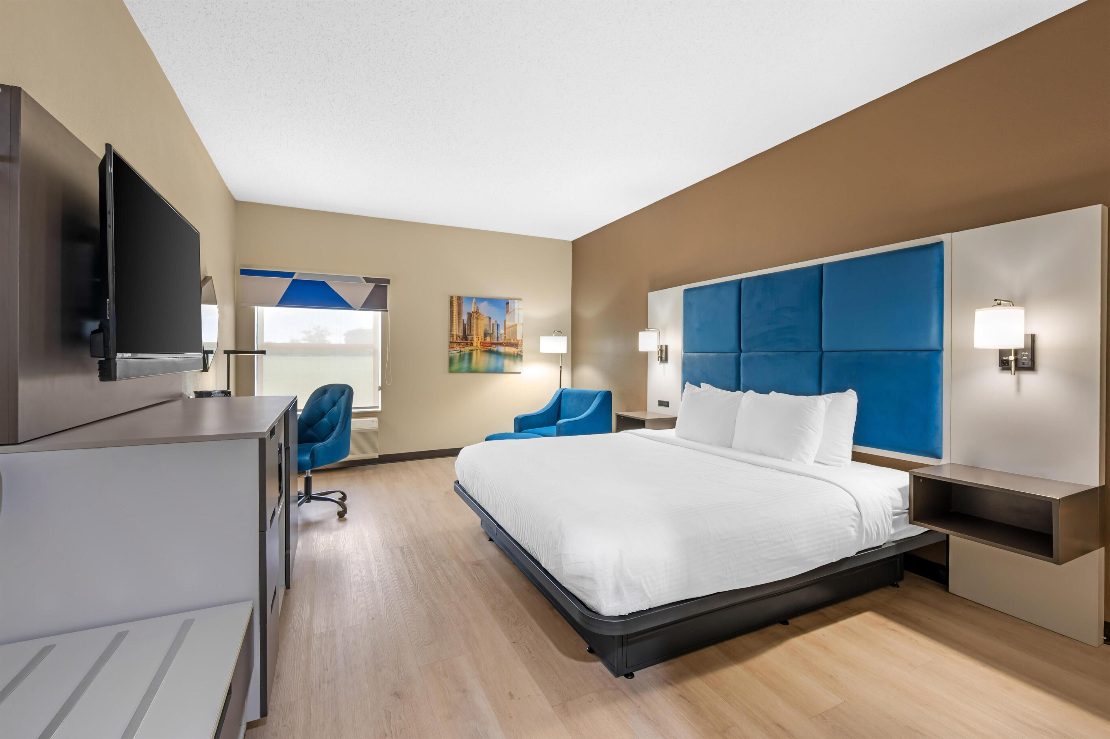 A hotel room with a bed, desk, chair, TV, artwork, and wall-mounted lights.
