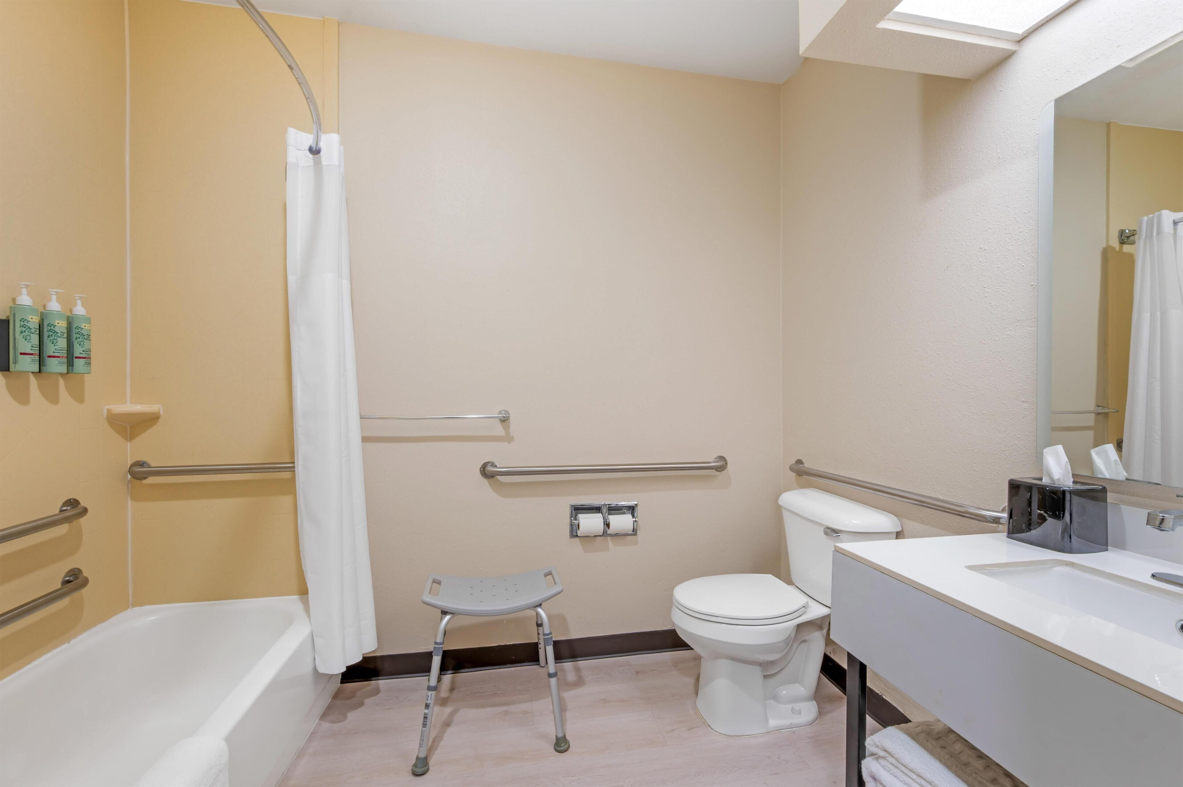 A bathroom with a bathtub, toilet, shower curtain, handrails, a step stool, and a mirror.