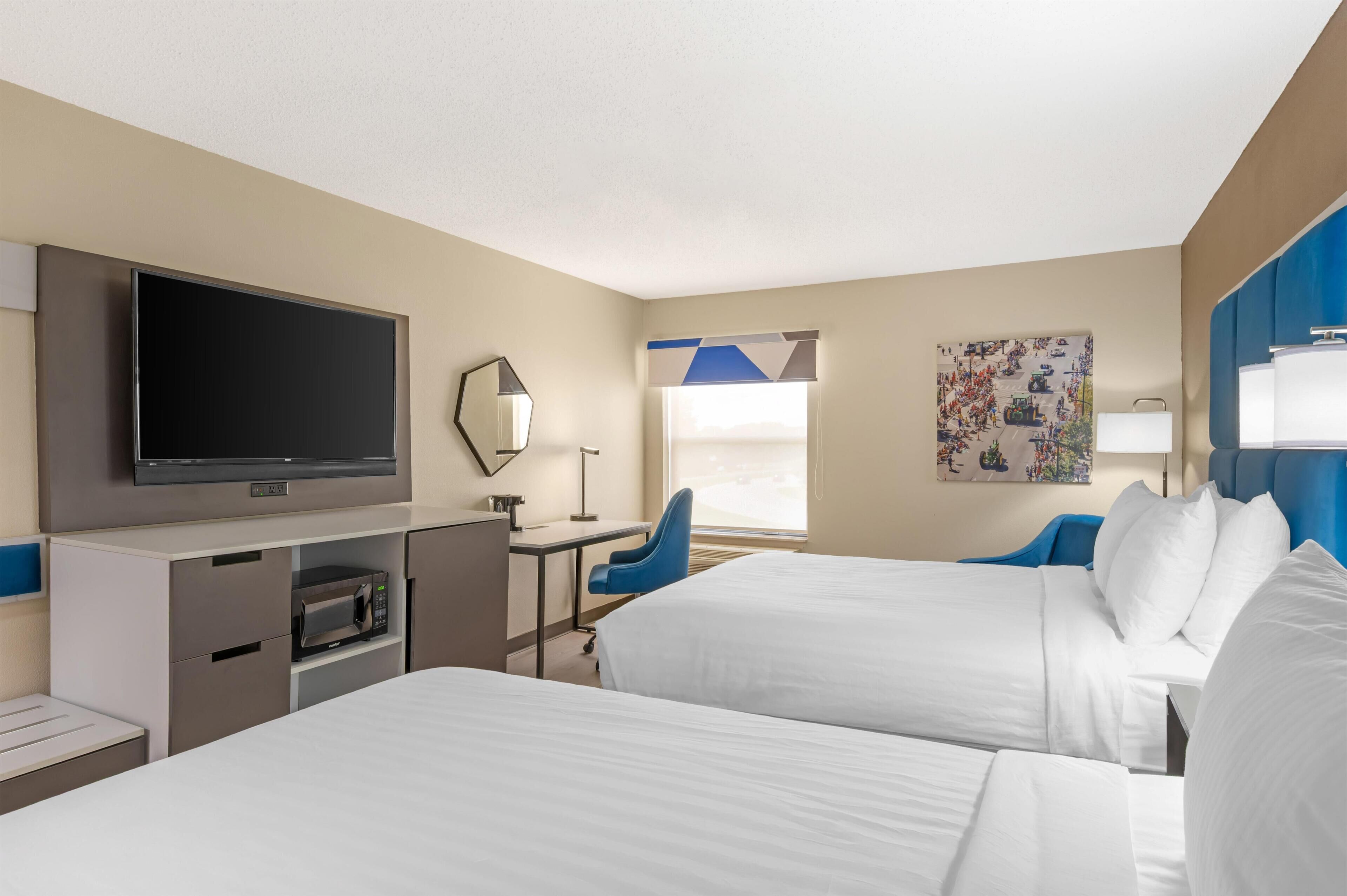 A hotel room with two beds, a TV, a desk with a chair, a microwave, and a window with a view.