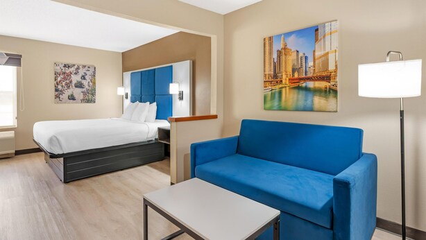A hotel room with a bed, a blue sofa, a small table, and a painting of a cityscape. - Best Western Forsyth - Decatur (Forsyth)
