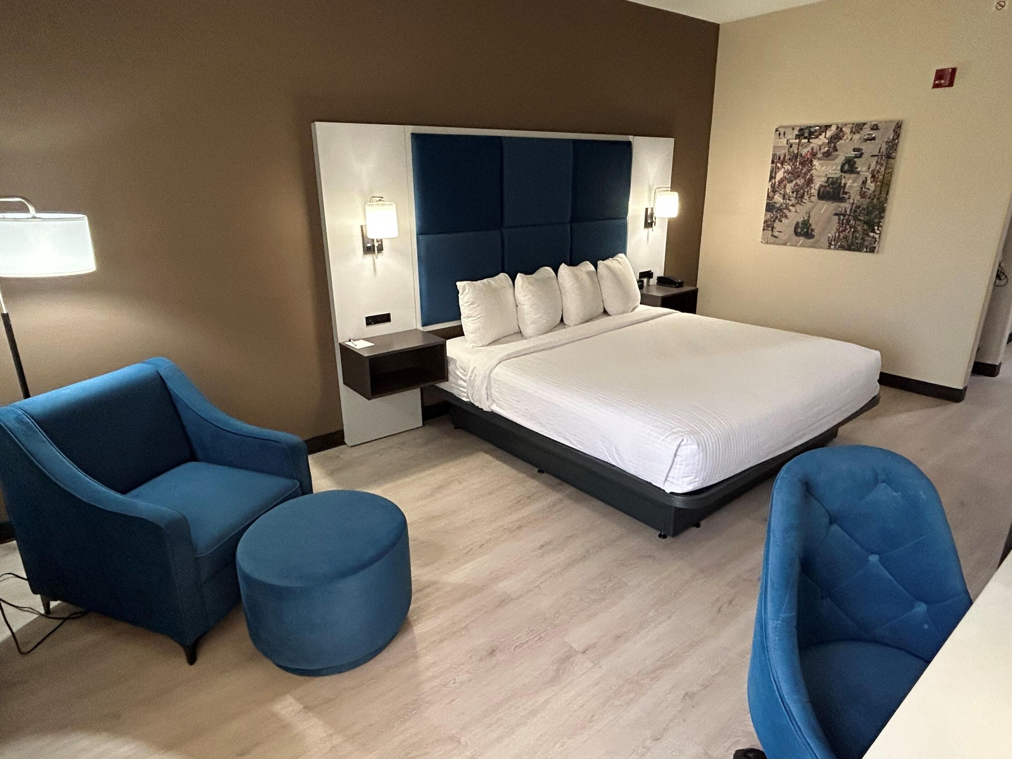 A hotel room with a large bed, two blue armchairs, a round ottoman, a nightstand with a lamp, and a wall-mounted artwork.