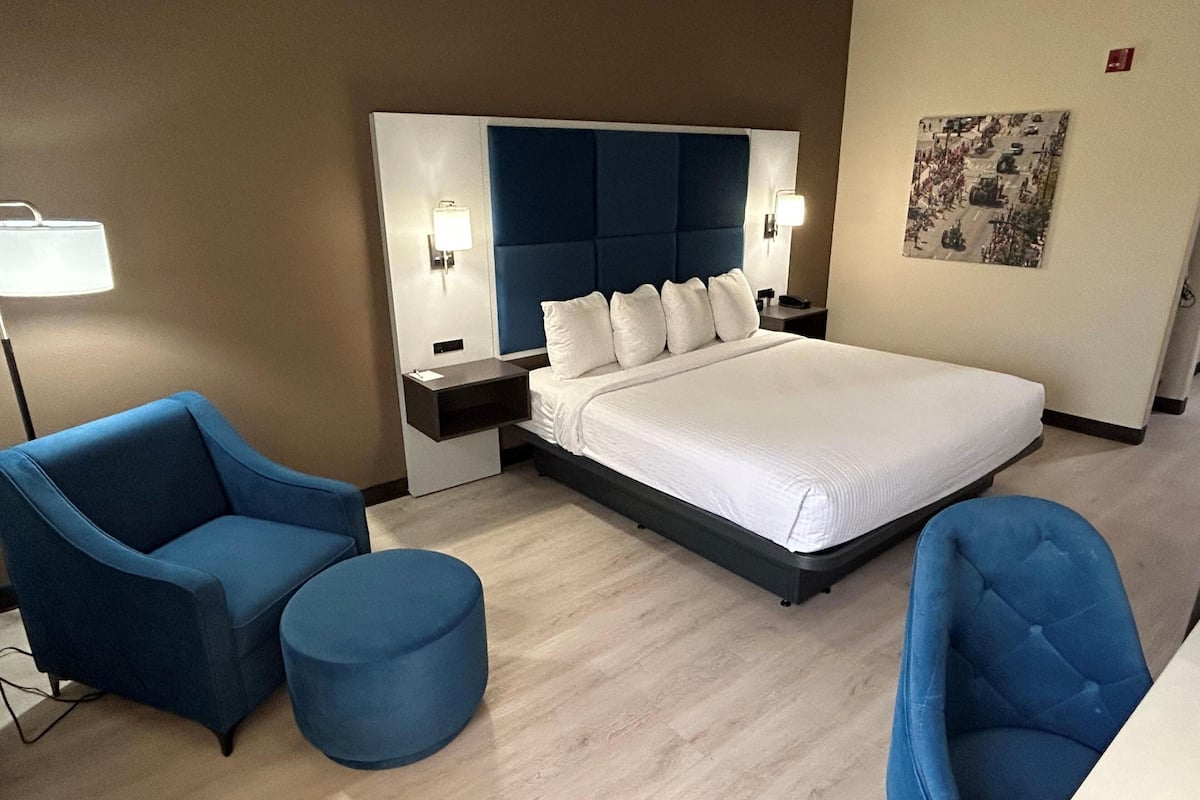 A hotel room with a large bed, two blue armchairs, a round ottoman, a nightstand with a lamp, and a wall-mounted artwork.