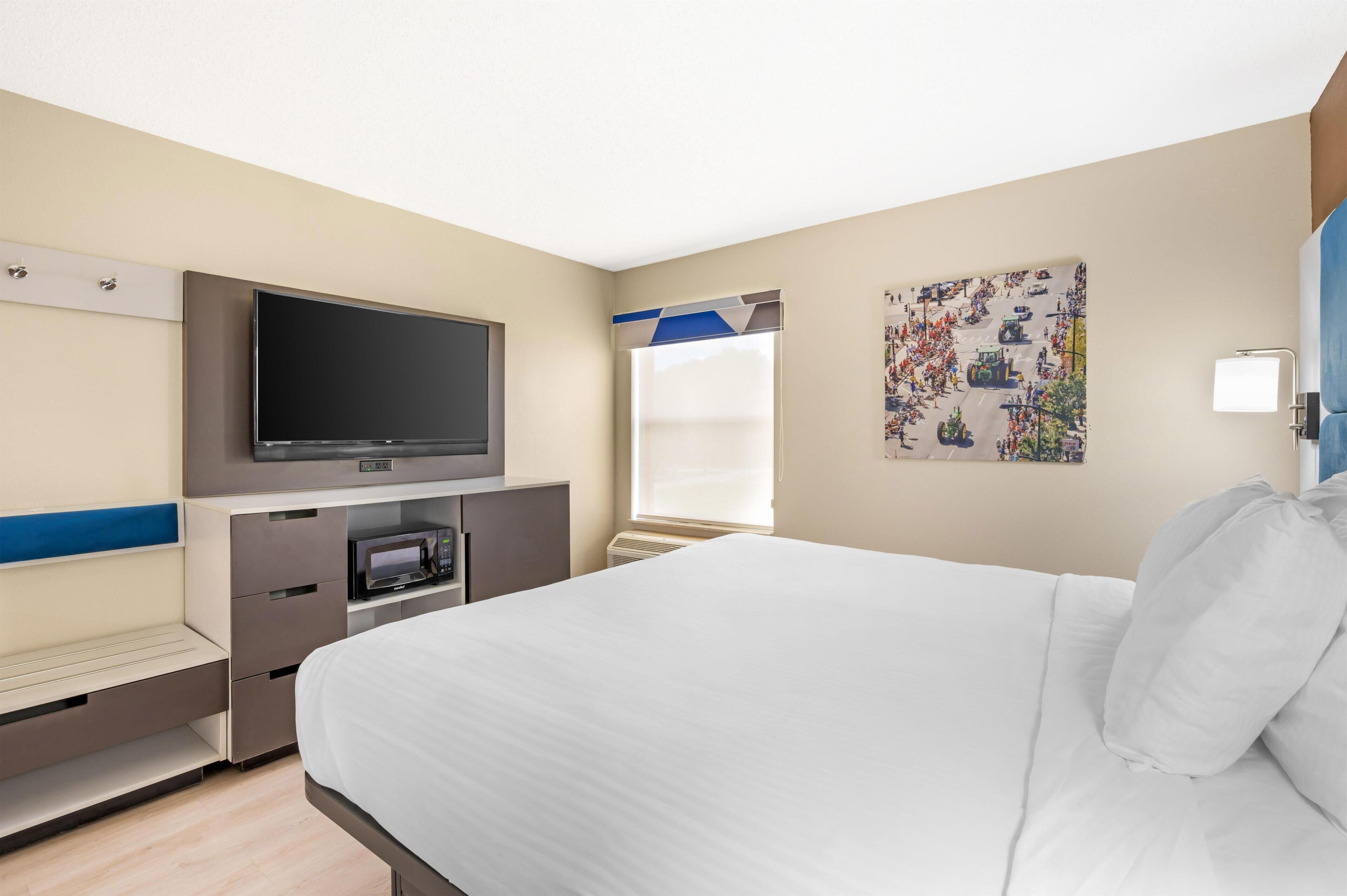 Habitación de hotel con cama, televisor de pantalla plana, microondas y ventana con persianas.