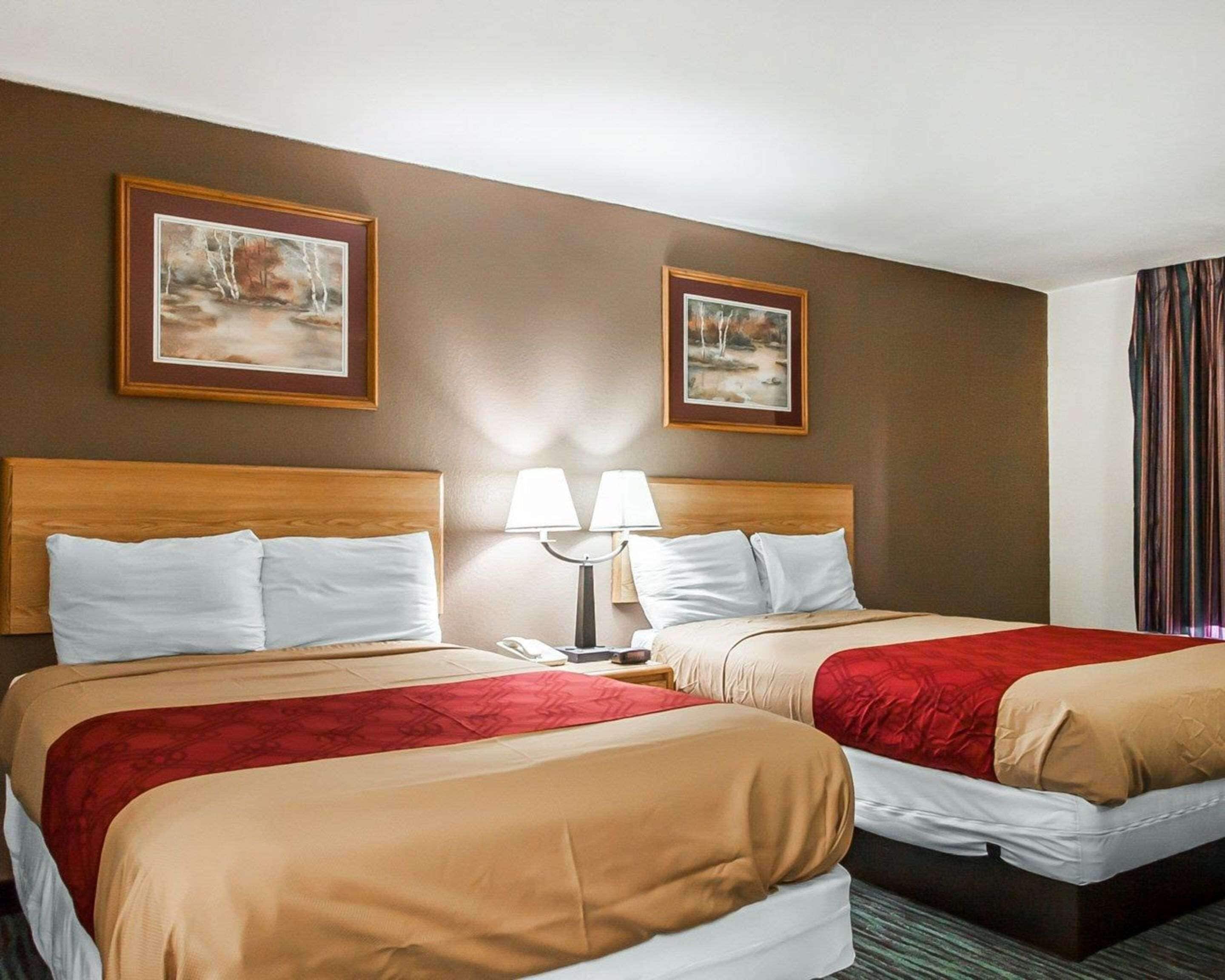 A hotel room with two beds, each with a wooden headboard, white bedding, and red throw blankets. There are two framed pictures on the wall, a nightstand with a lamp, and a striped curtain.