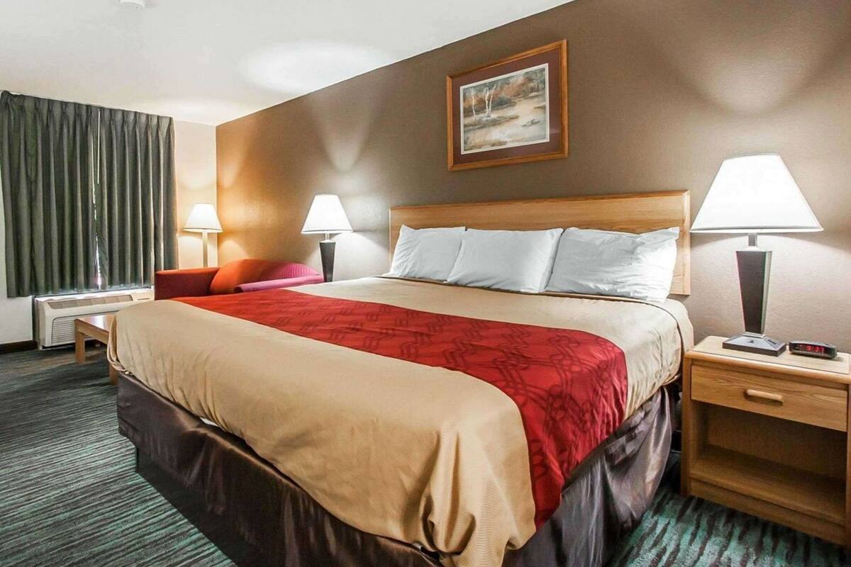 A hotel room with a bed, two bedside tables, a nightstand, a lamp, a painting on the wall, and a window with curtains.