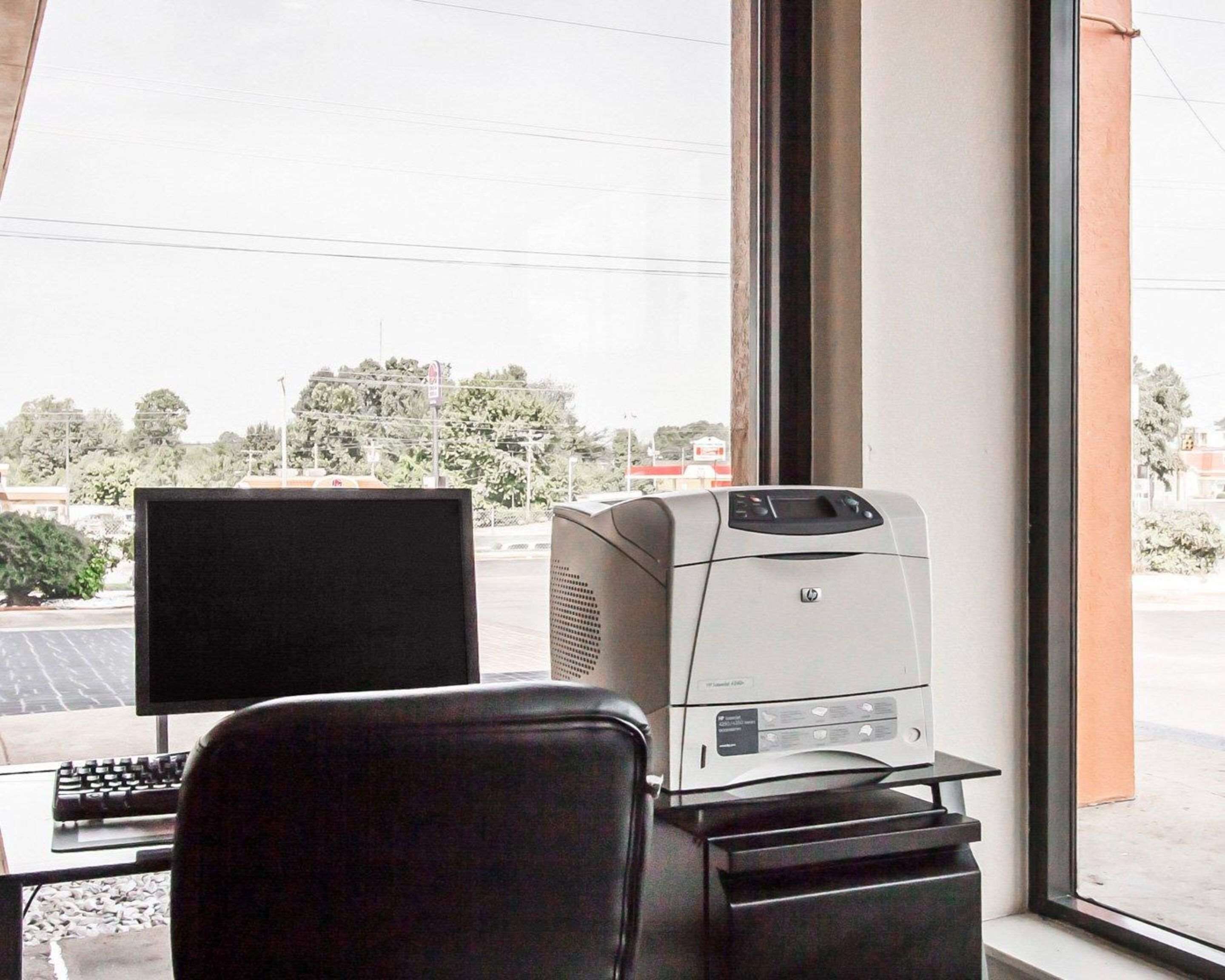 A workspace with a computer, printer, and chair near a window with a view of a parking lot and trees.
