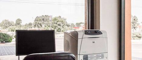 A workspace with a computer, printer, and chair near a window with a view of a parking lot and trees.