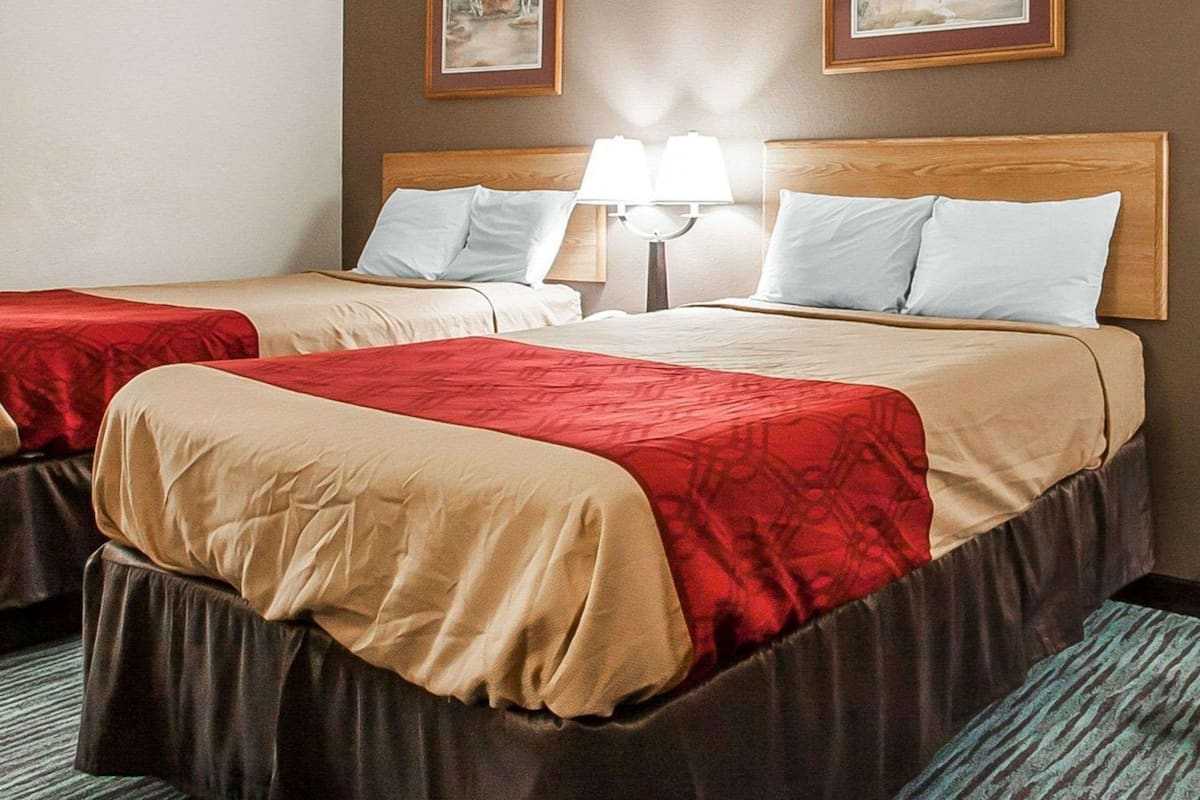 A hotel room with two beds, each with a red and beige bedspread, a wooden headboard, and a nightstand with a lamp.