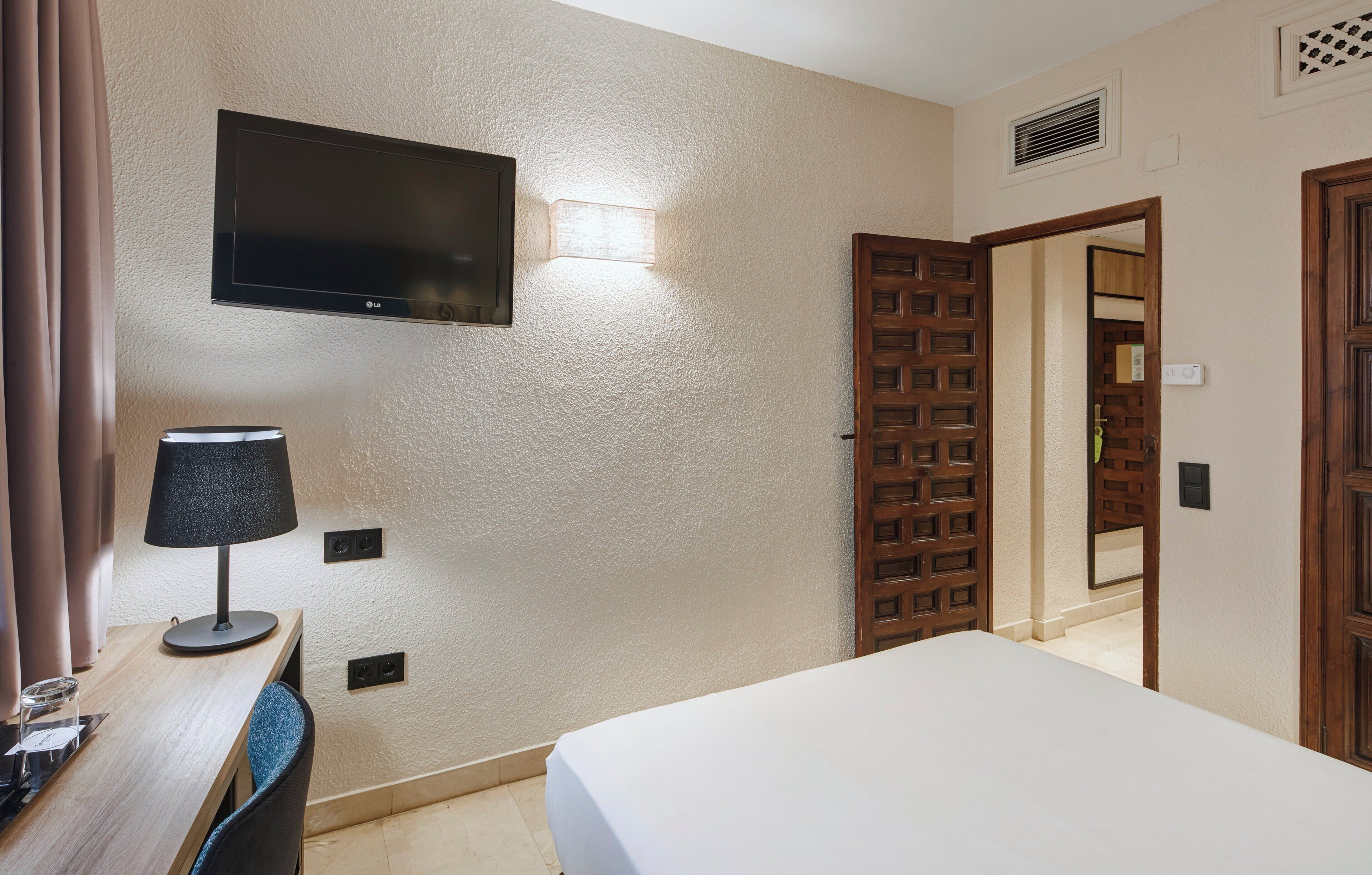 A hotel room with a bed, a desk with a lamp, a television mounted on the wall, and a wooden door.