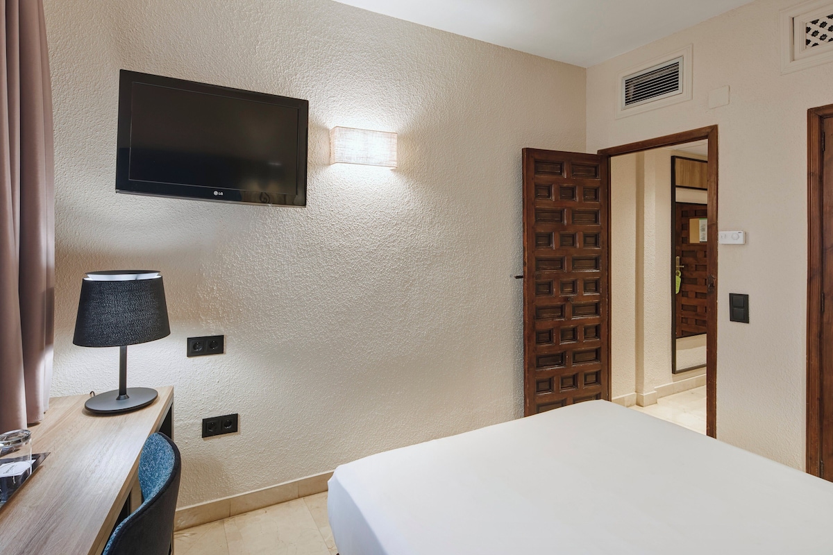 A hotel room with a bed, a desk with a lamp, a television mounted on the wall, and a wooden door.