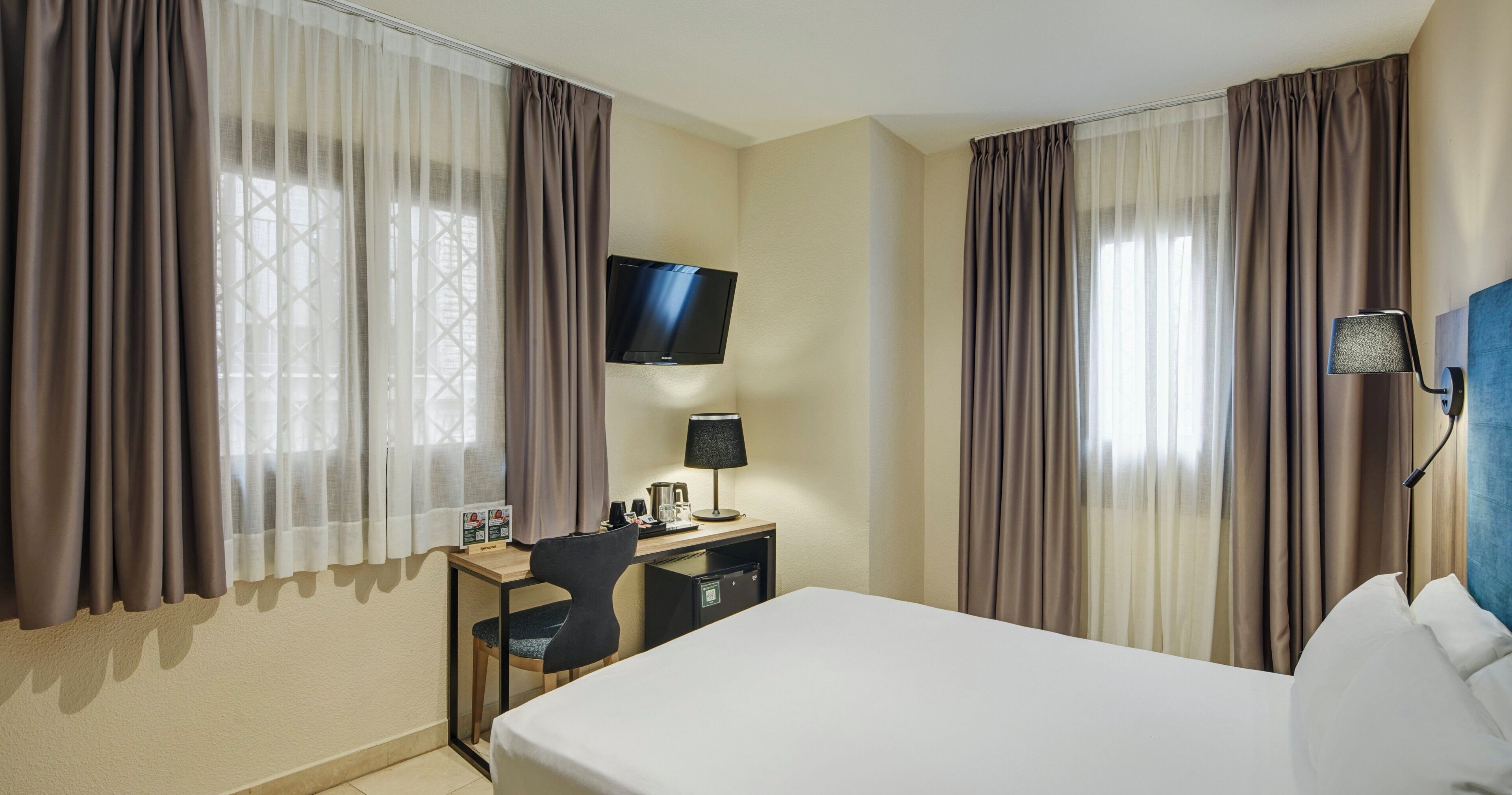 Una camera d'albergo con un letto, una scrivania, una sedia, televisione, lampada e tende.