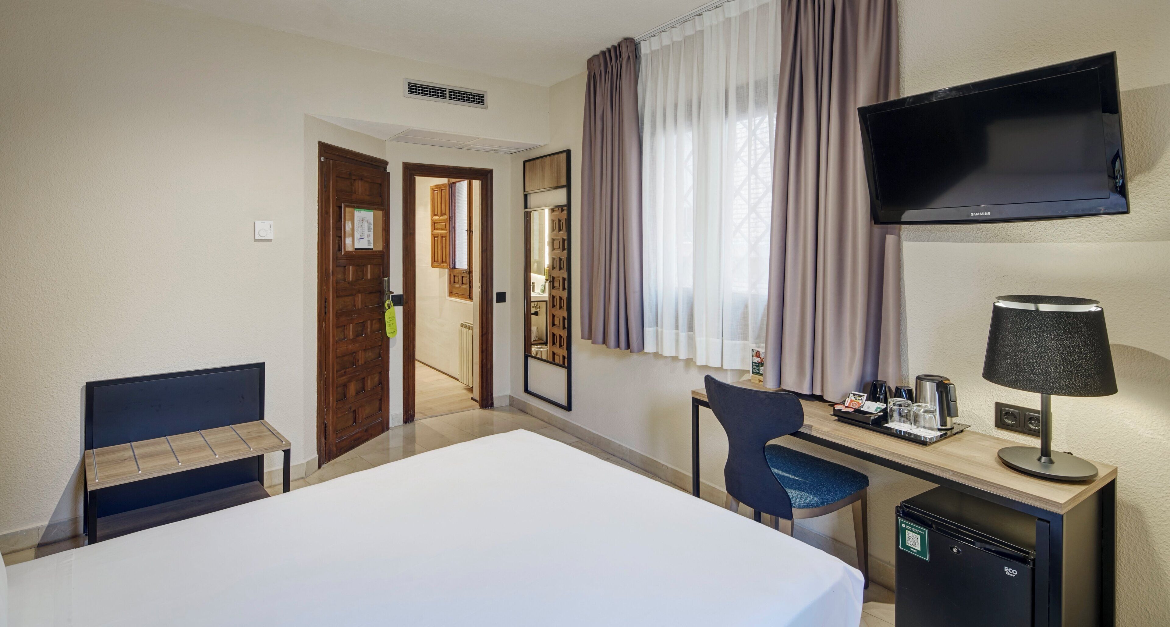 A hotel room with a bed, a desk with a chair, a television, and a minibar.