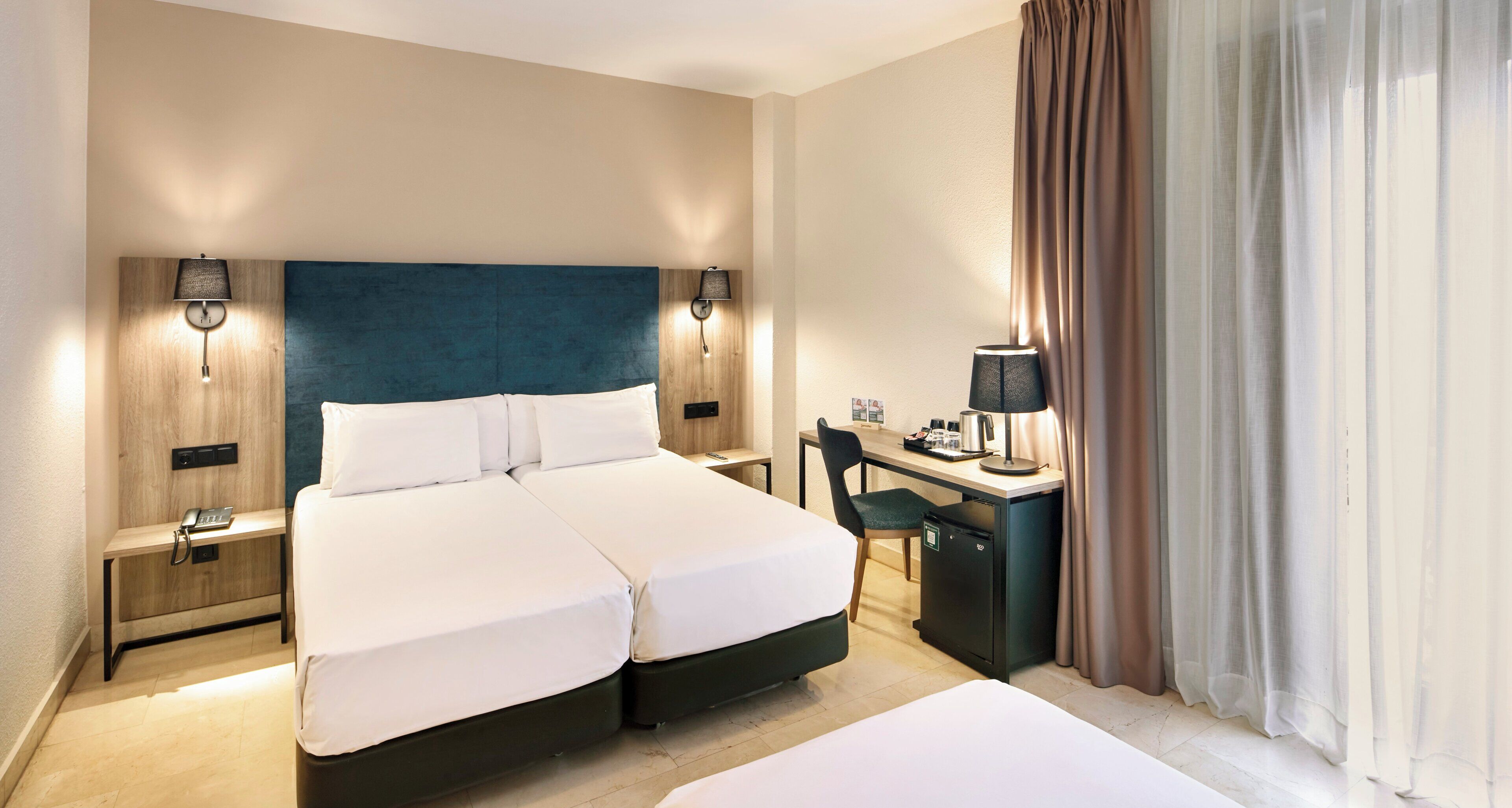 A hotel room with a double bed, a desk with a chair, a small refrigerator, and two bedside tables with lamps.