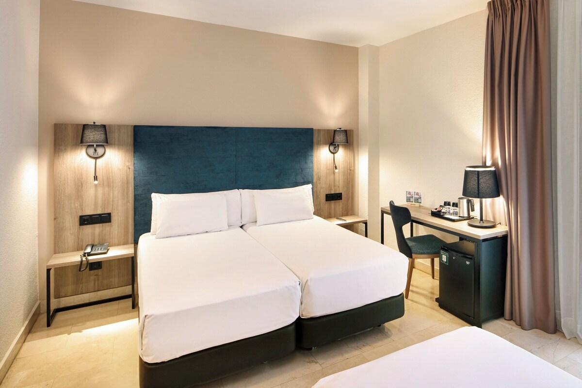 A hotel room with a double bed, a desk with a chair, a small refrigerator, and two bedside tables with lamps.