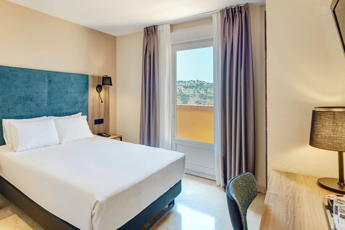 Ein Hotelzimmer mit einem Bett, zwei Nachttischen, einer Lampe, einem Sessel und Blick ins Freie.