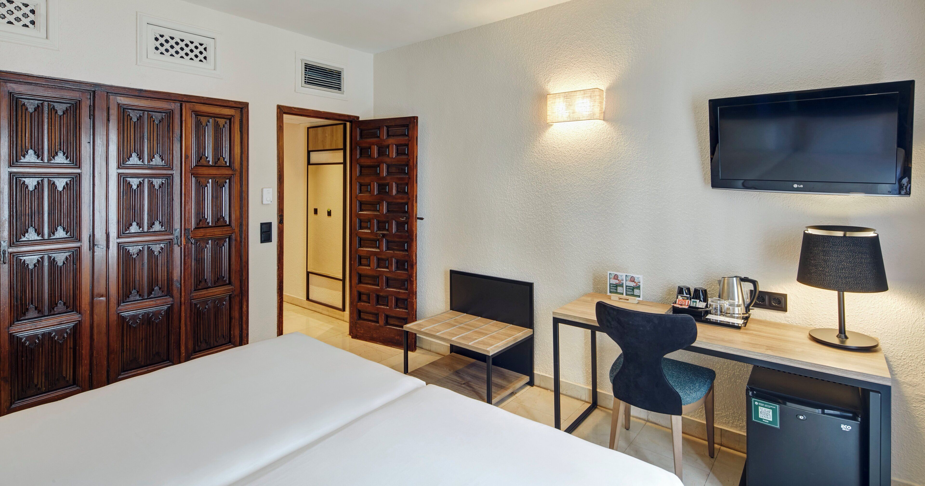A hotel room with a bed, desk, chair, TV, and a minibar.