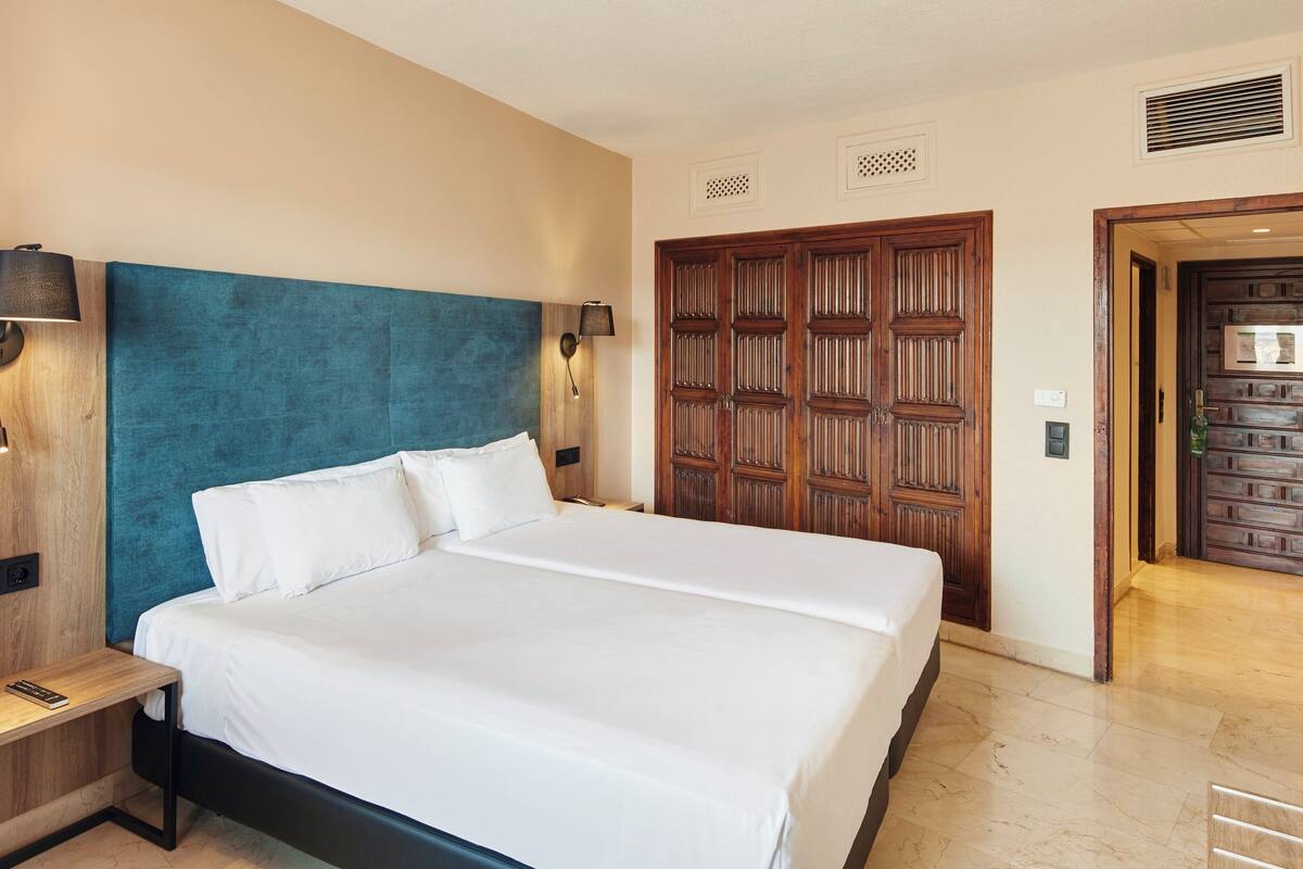 Une chambre d’hôtel avec un grand lit, une tête de lit en bois, deux lampes fixées au mur, une table de chevet et une porte ornée d’un panneau décoratif.