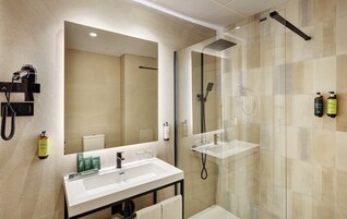 A modern bathroom with a large mirror, a white sink, a handheld showerhead, and two bottles of shampoo.