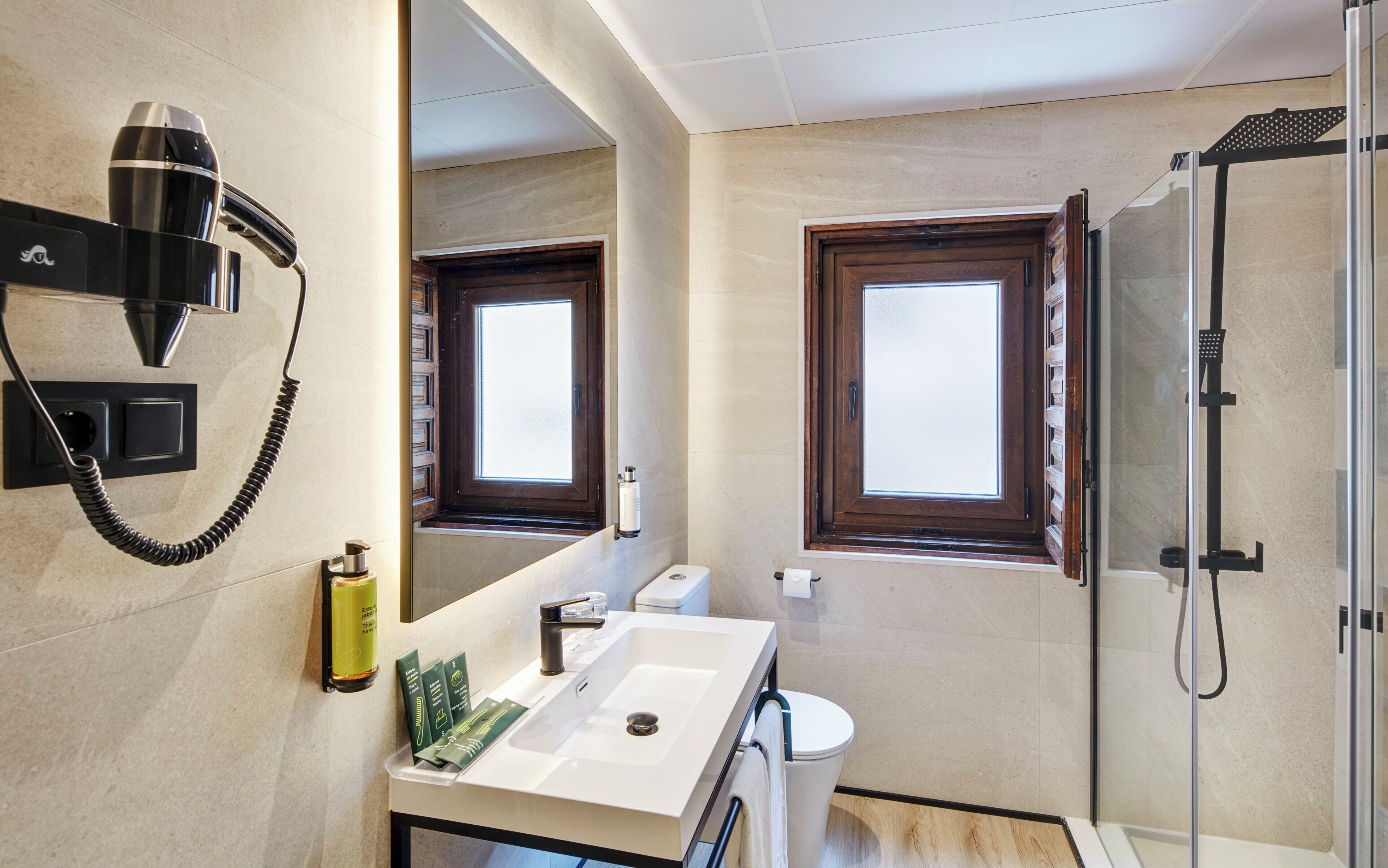 A modern bathroom with a toilet, sink, and shower. There is a hairdryer mounted on the wall, a bottle of shampoo, and a towel rack.